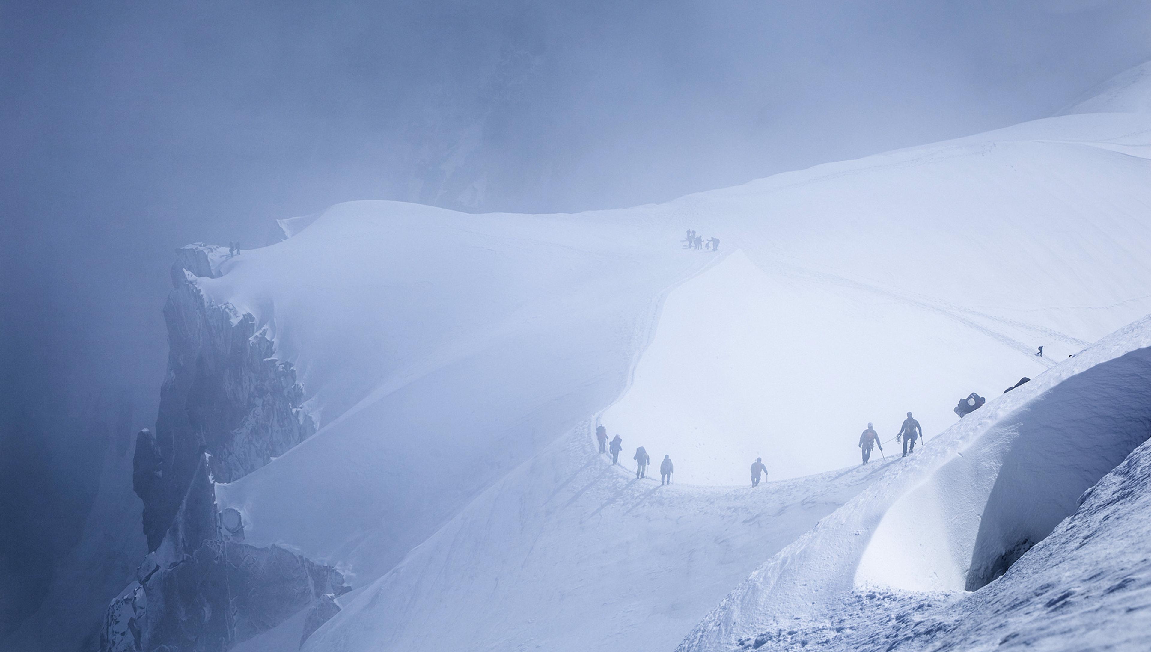 Several climbers trek across a snow-covered mountain ridge, enshrouded in mist, with rocky outcrops visible to the left. The path they follow is narrow and steep, demonstrating the challenging conditions of high-altitude mountaineering.