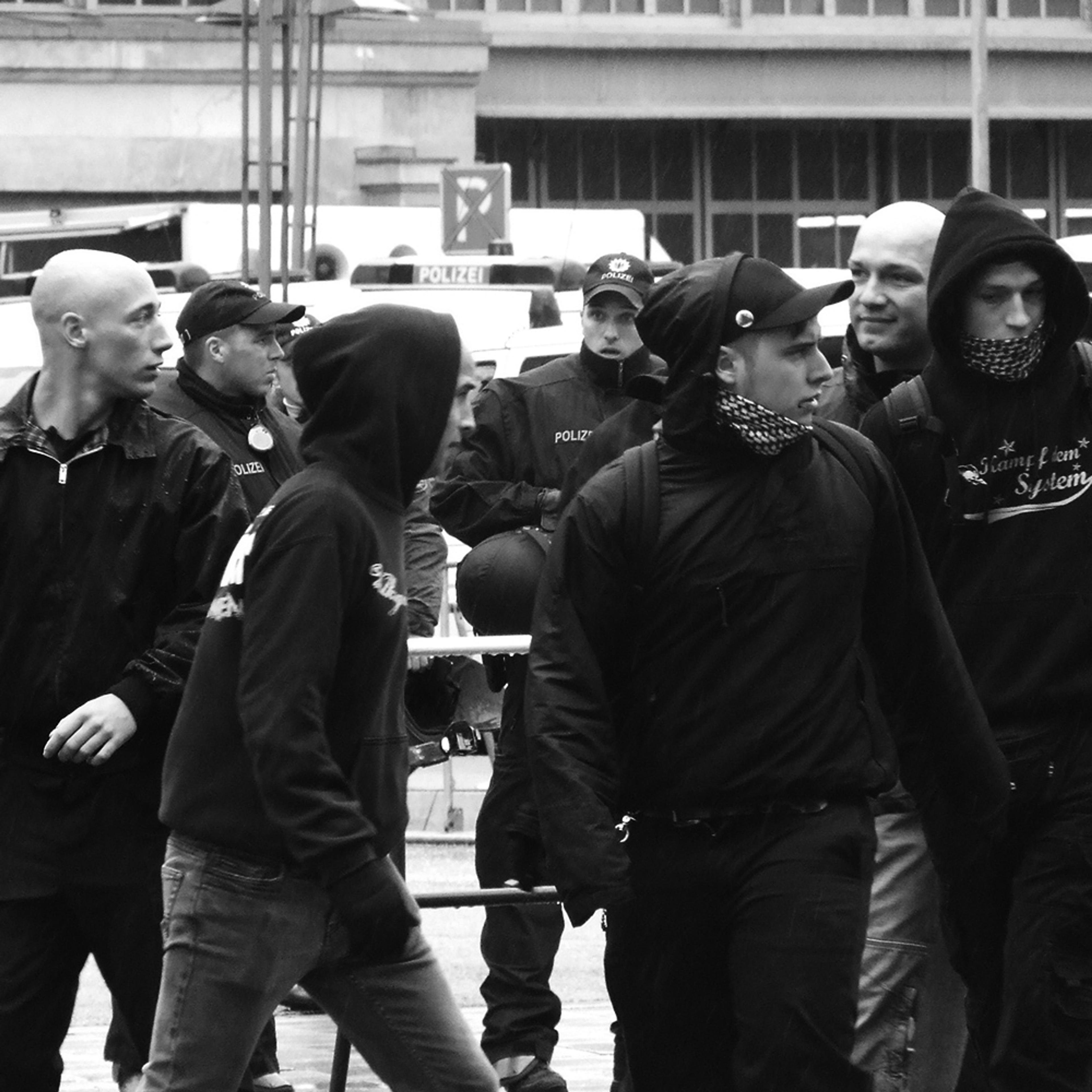 Black and white photo of white young men in dark clothes, some hooded, with police officers and police vans in the background.