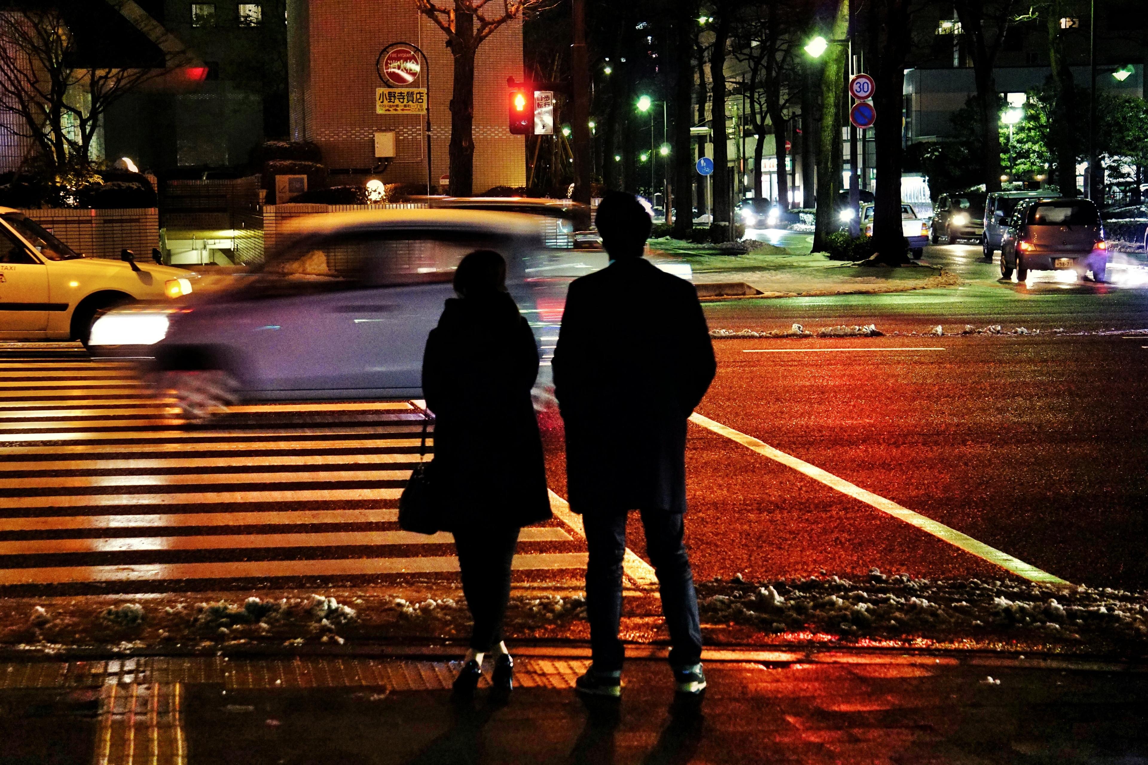 Photo of two people standing at a zebra crossing at night, with cars passing by and illuminated streetlights.