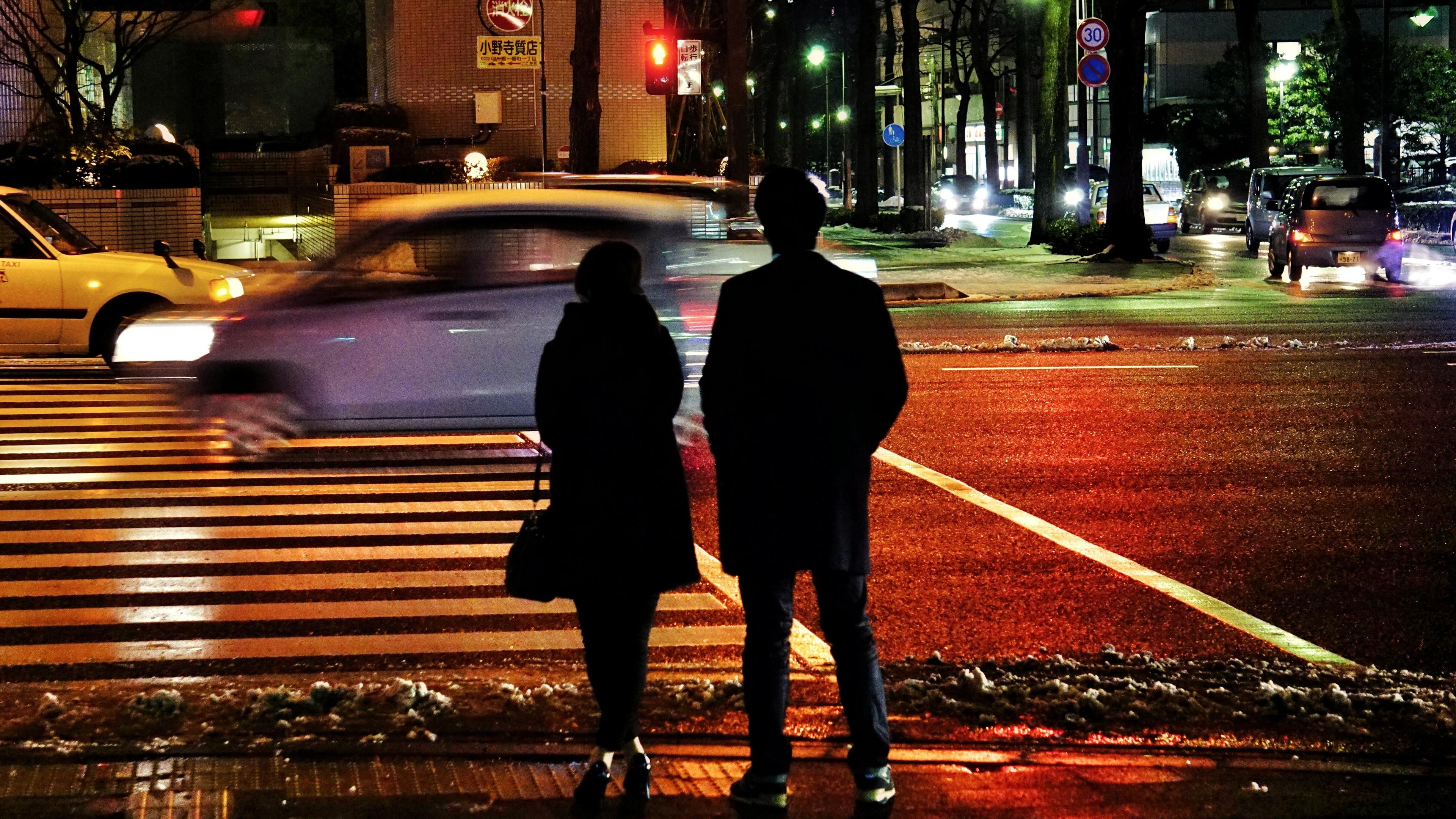 Photo of two people standing at a zebra crossing at night, with cars passing by and illuminated streetlights.