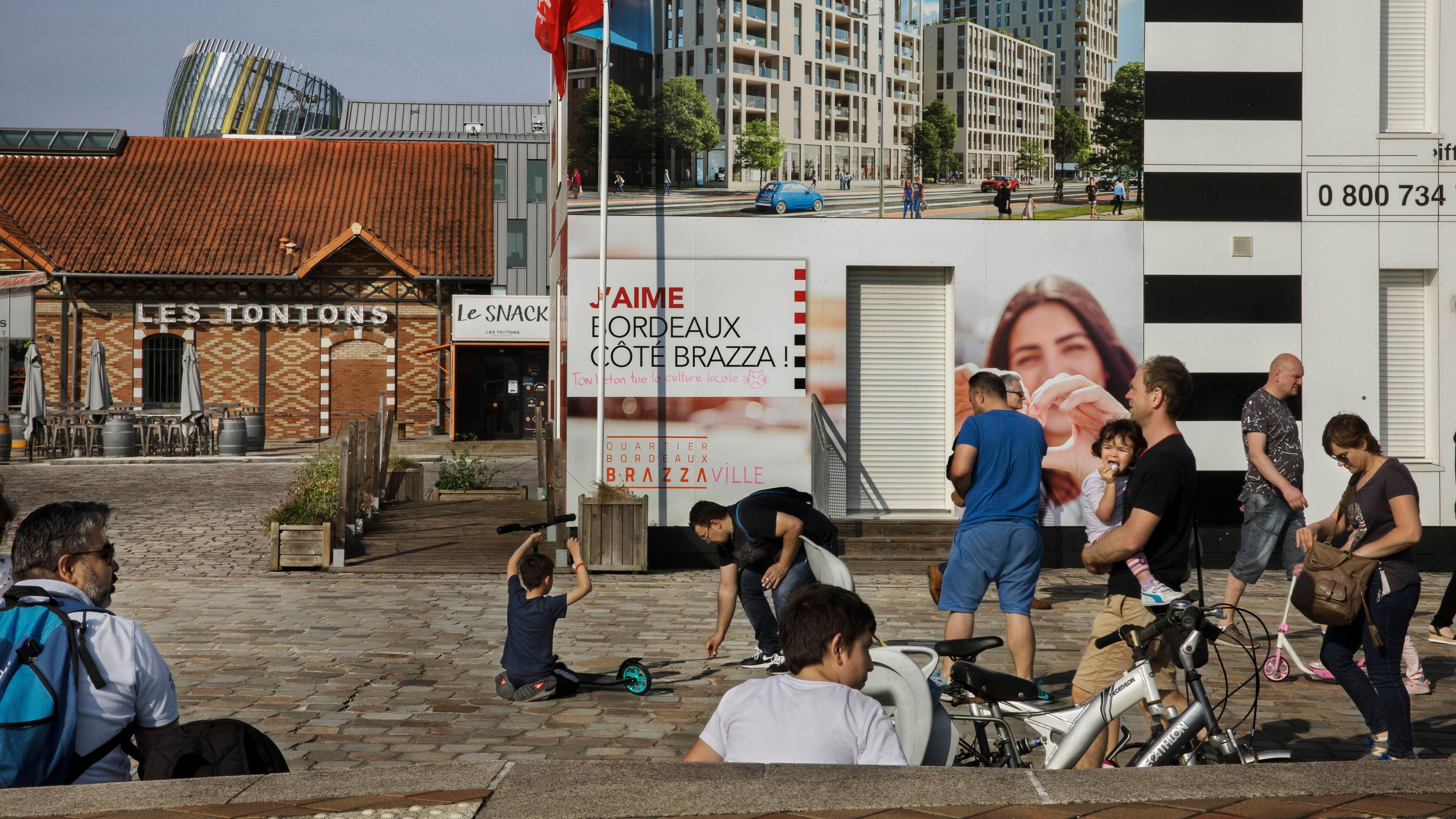 Photo of people in a sunny square with bikes and scooters, near Les Tontons and a large building advertisement in Bordeaux.