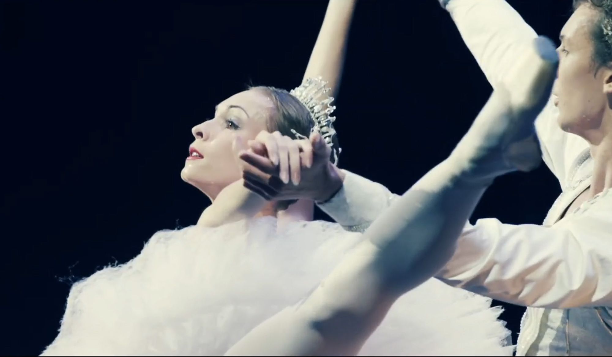 Two ballet dancers performing an elegant move, the ballerina in a white tutu with an outstretched leg and arms entwined with her partner.