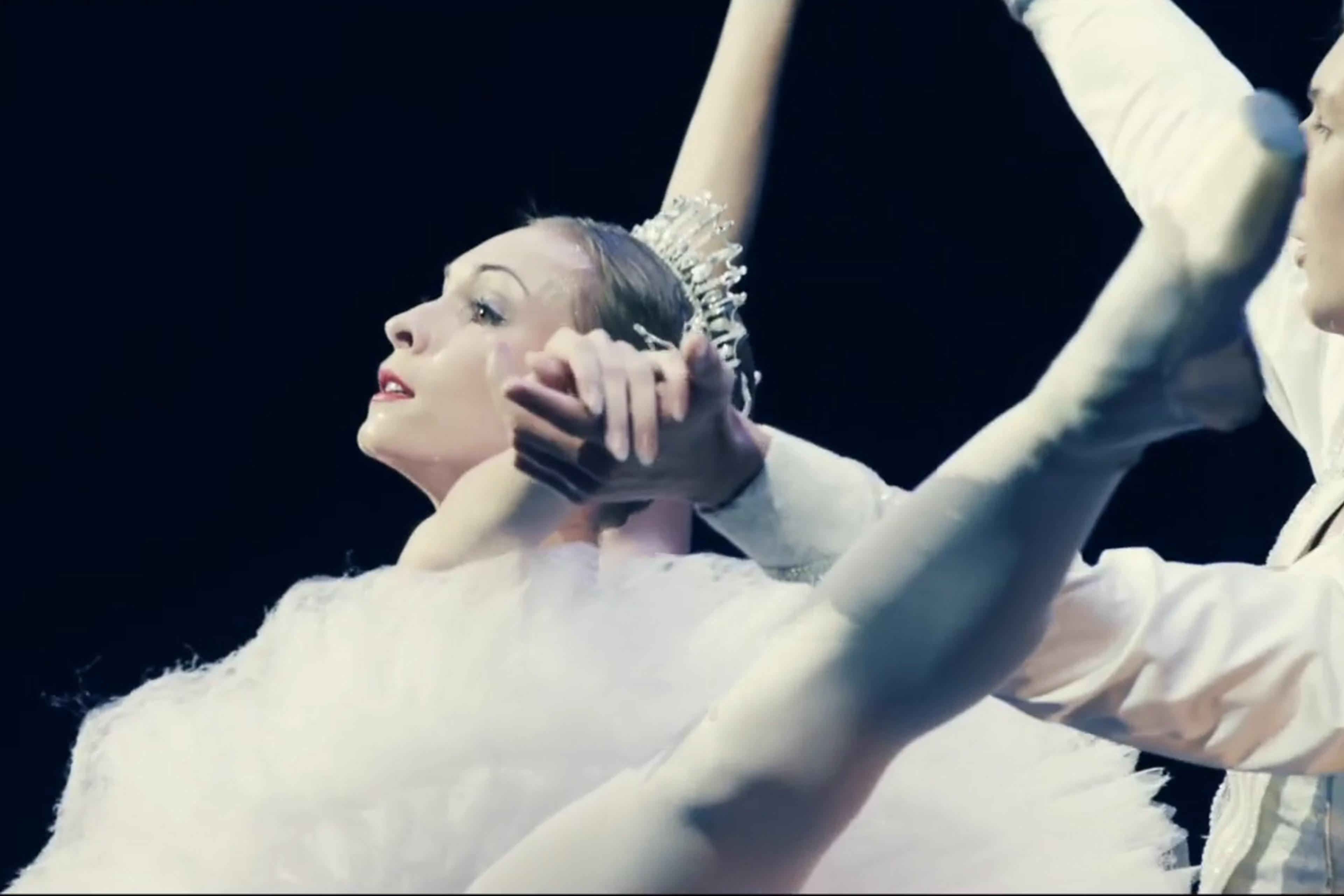 Two ballet dancers performing an elegant move, the ballerina in a white tutu with an outstretched leg and arms entwined with her partner.