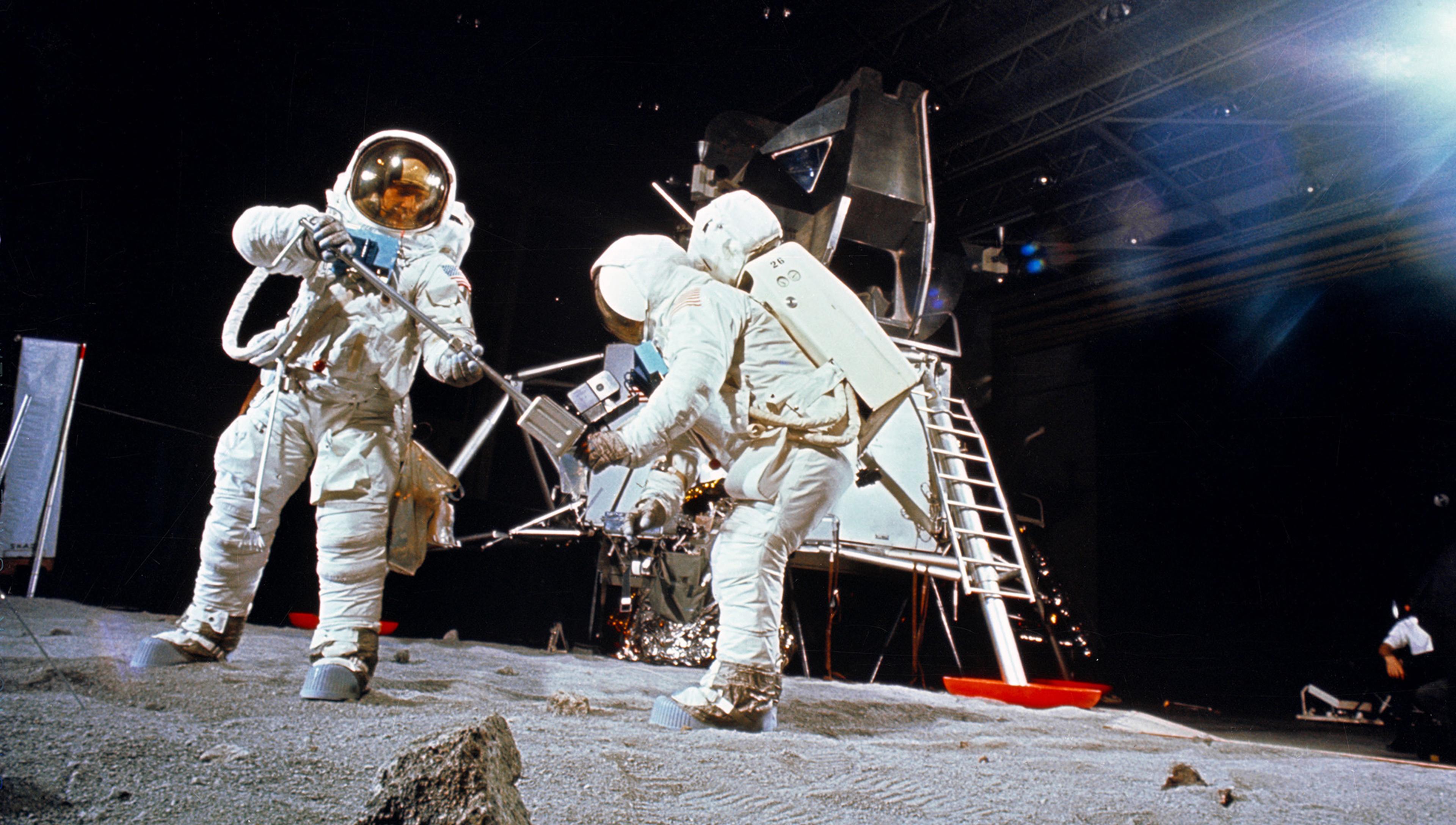 Photo of two astronauts in spacesuits on a simulated lunar surface with a lunar module in the background.