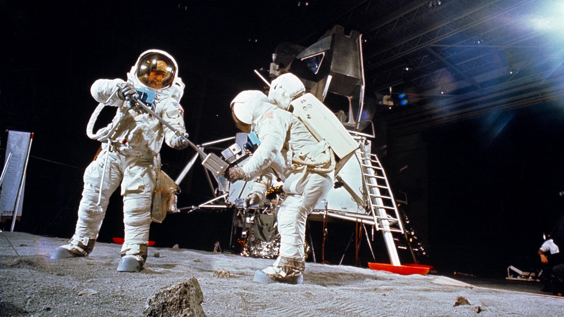Photo of two astronauts in spacesuits on a simulated lunar surface with a lunar module in the background.