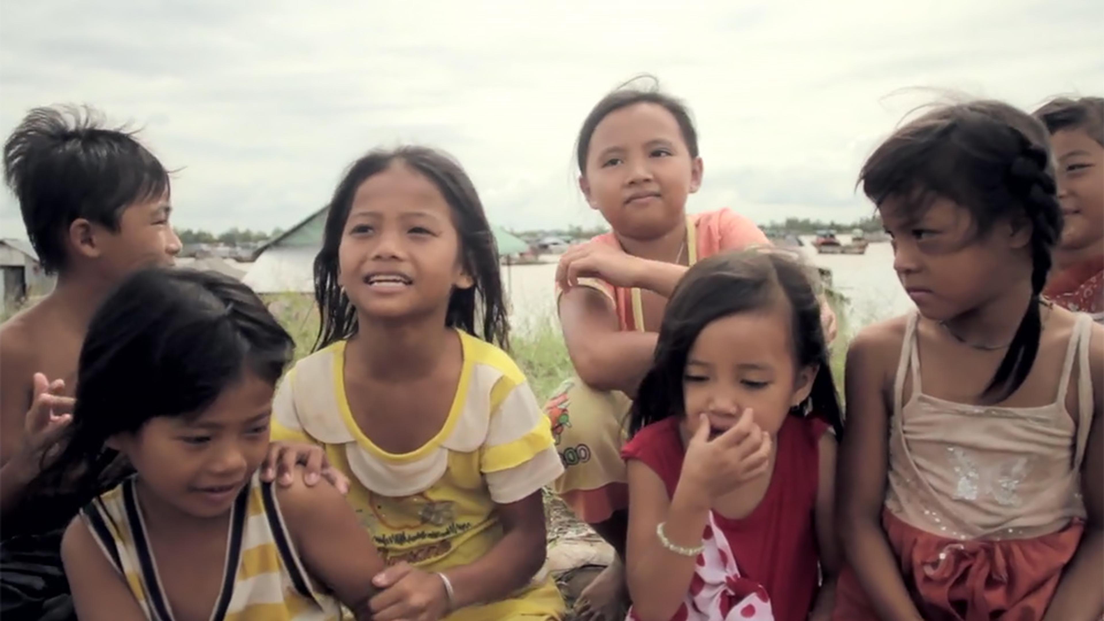 Seven children smiling and interacting outdoors with a blurred background of boats and houses on the water.