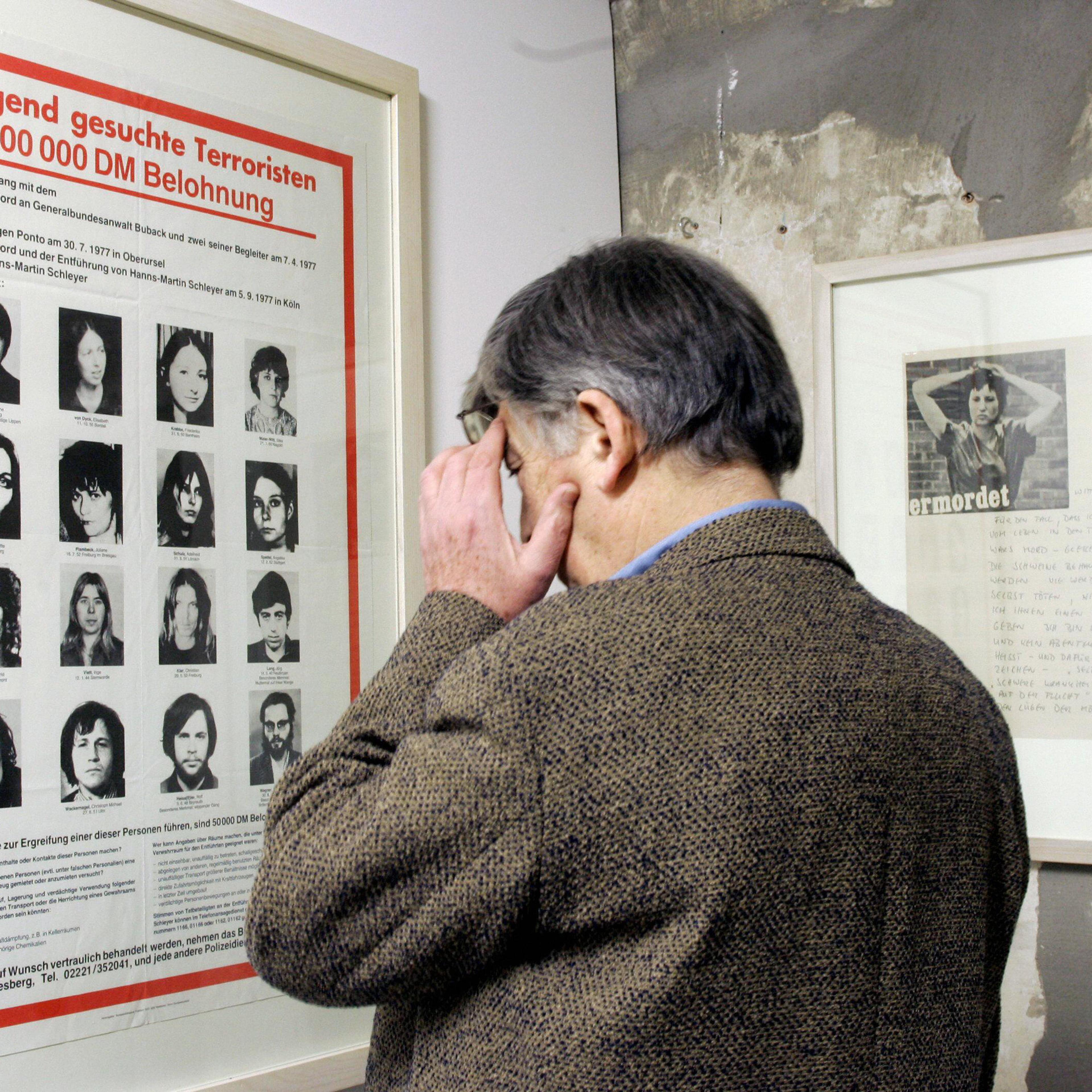 A man in a tweed jacket viewing a framed German wanted poster on a wall in a museum or gallery setting.