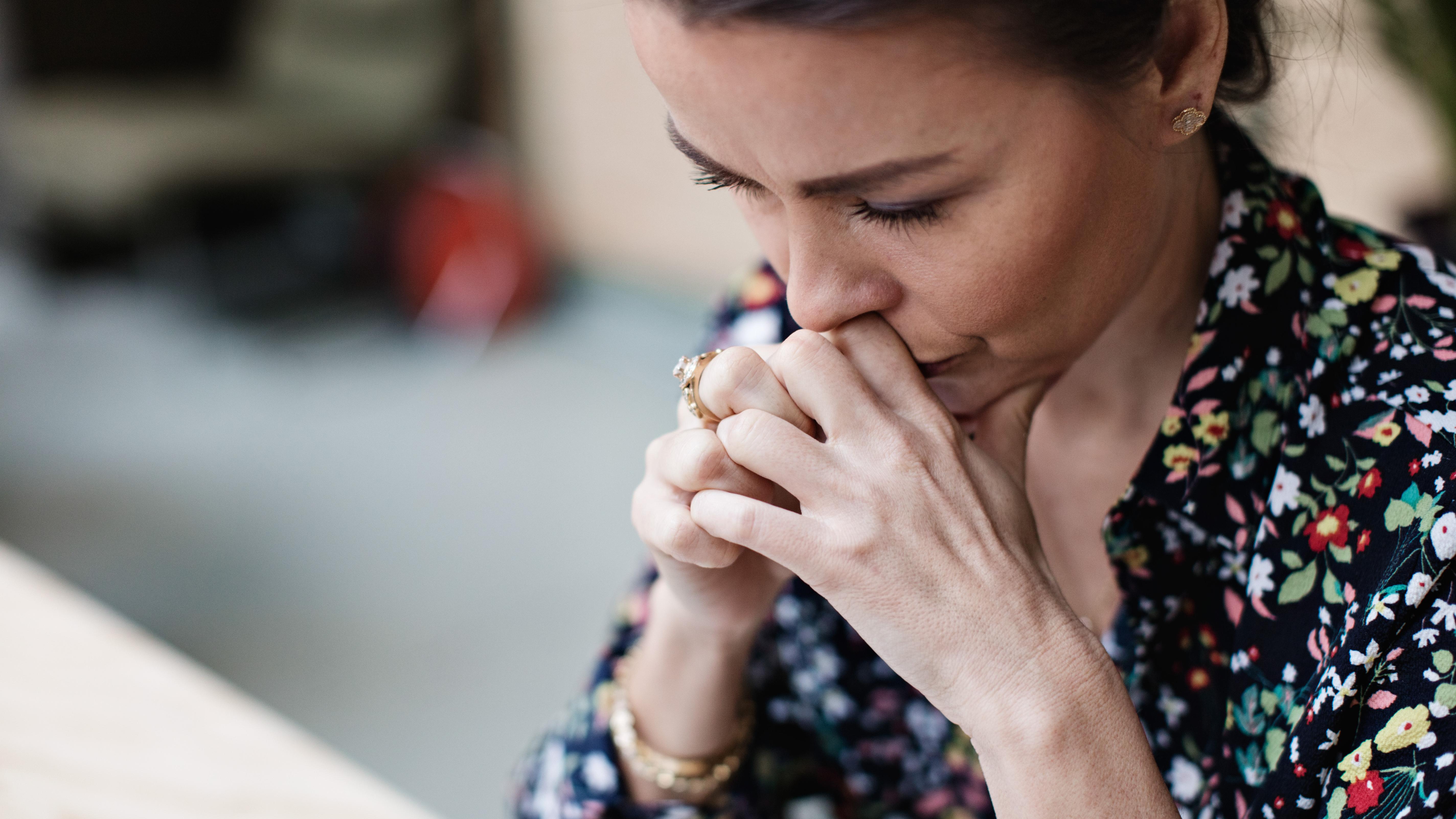 Photo of a woman in a floral shirt, appearing deep in thought with her hands clasped near her face.