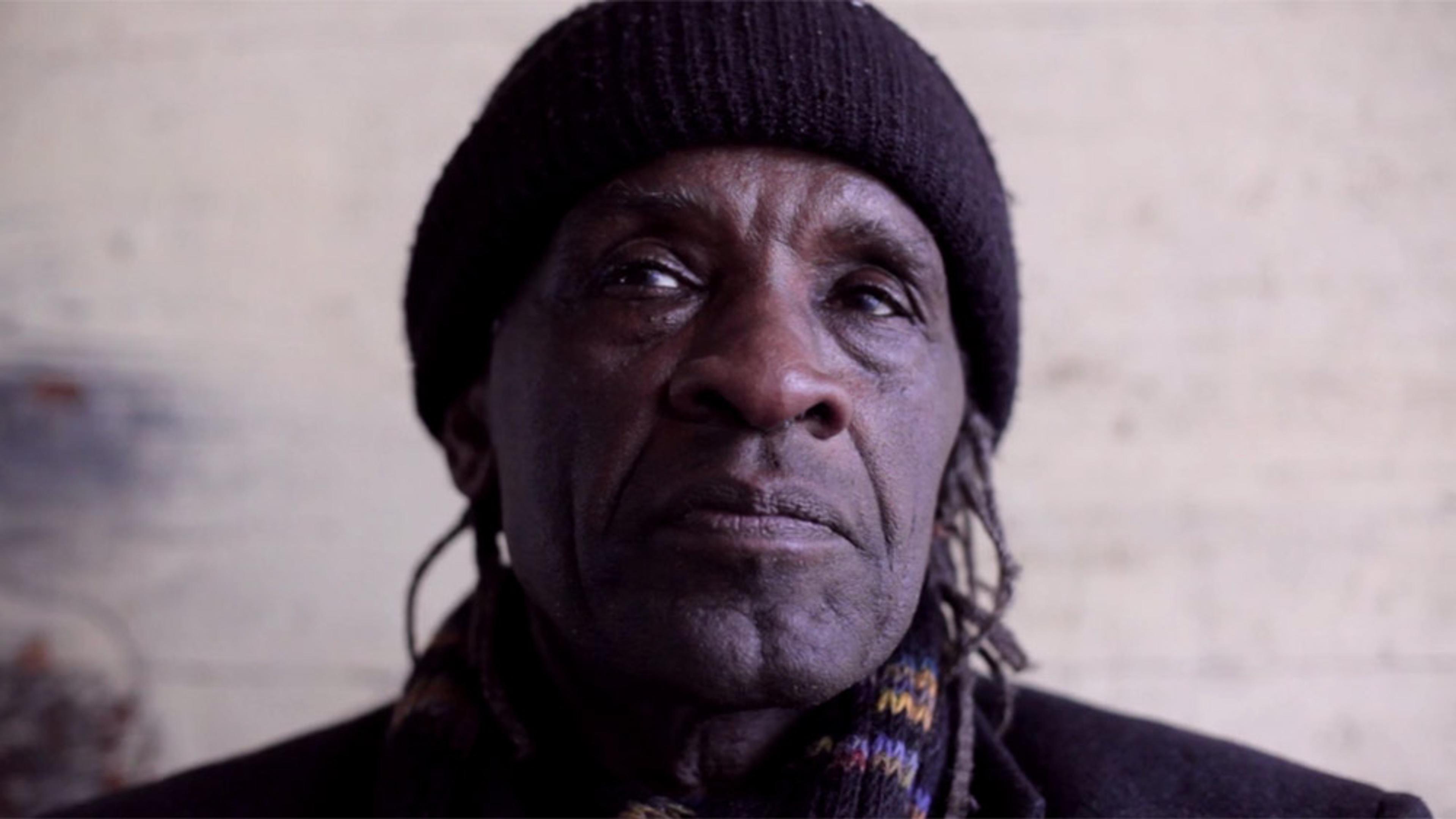 An elderly man wearing a black beanie and scarf, looking forward with a neutral expression, set against a blurred background.