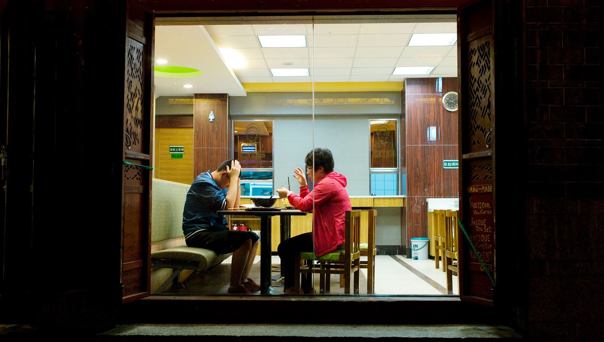 Two people facing each other in a restaurant, one gesticulates, the other has head in his hands, seen through a window at night.