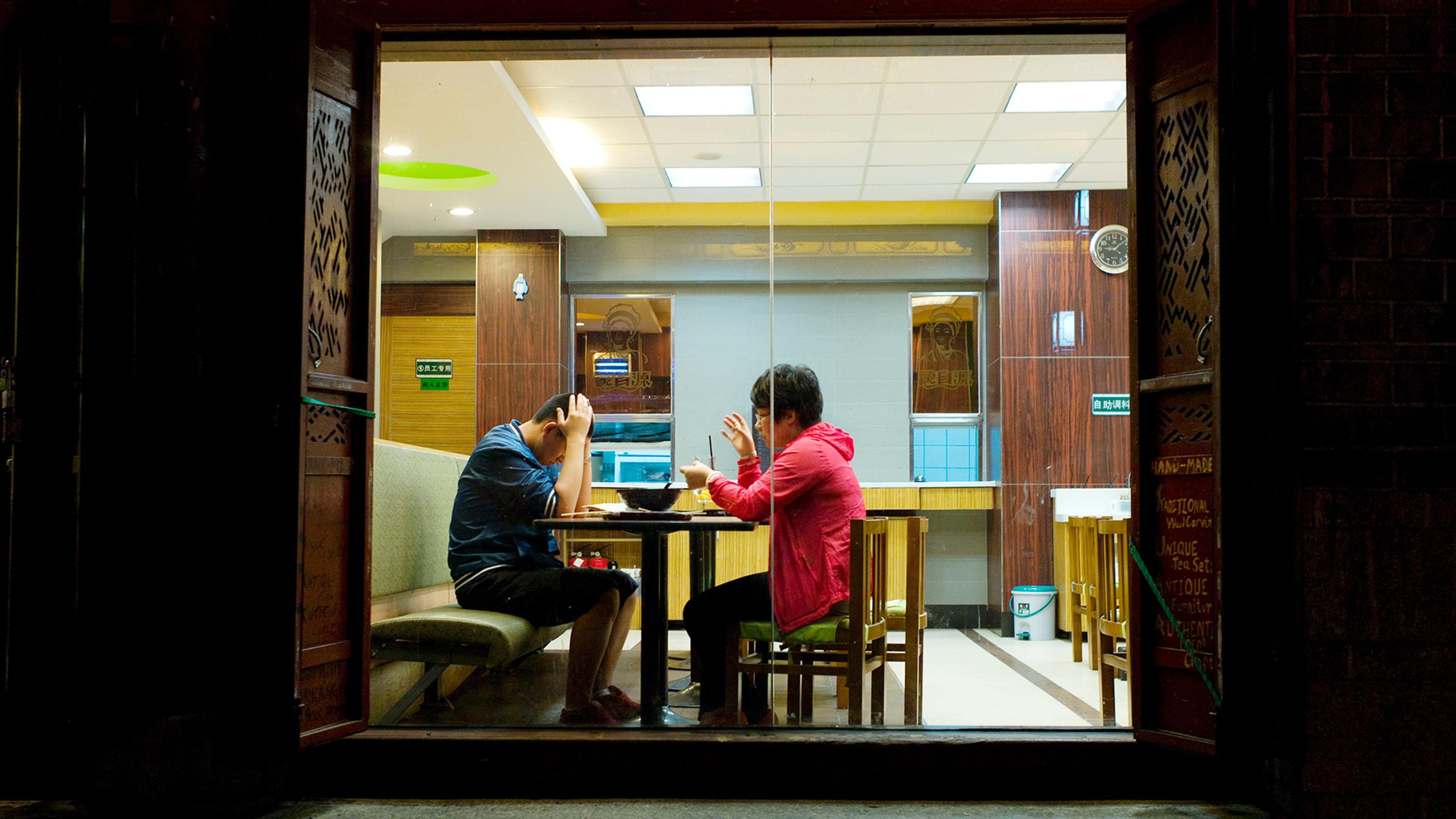 Two people facing each other in a restaurant, one gesticulates, the other has head in his hands, seen through a window at night.