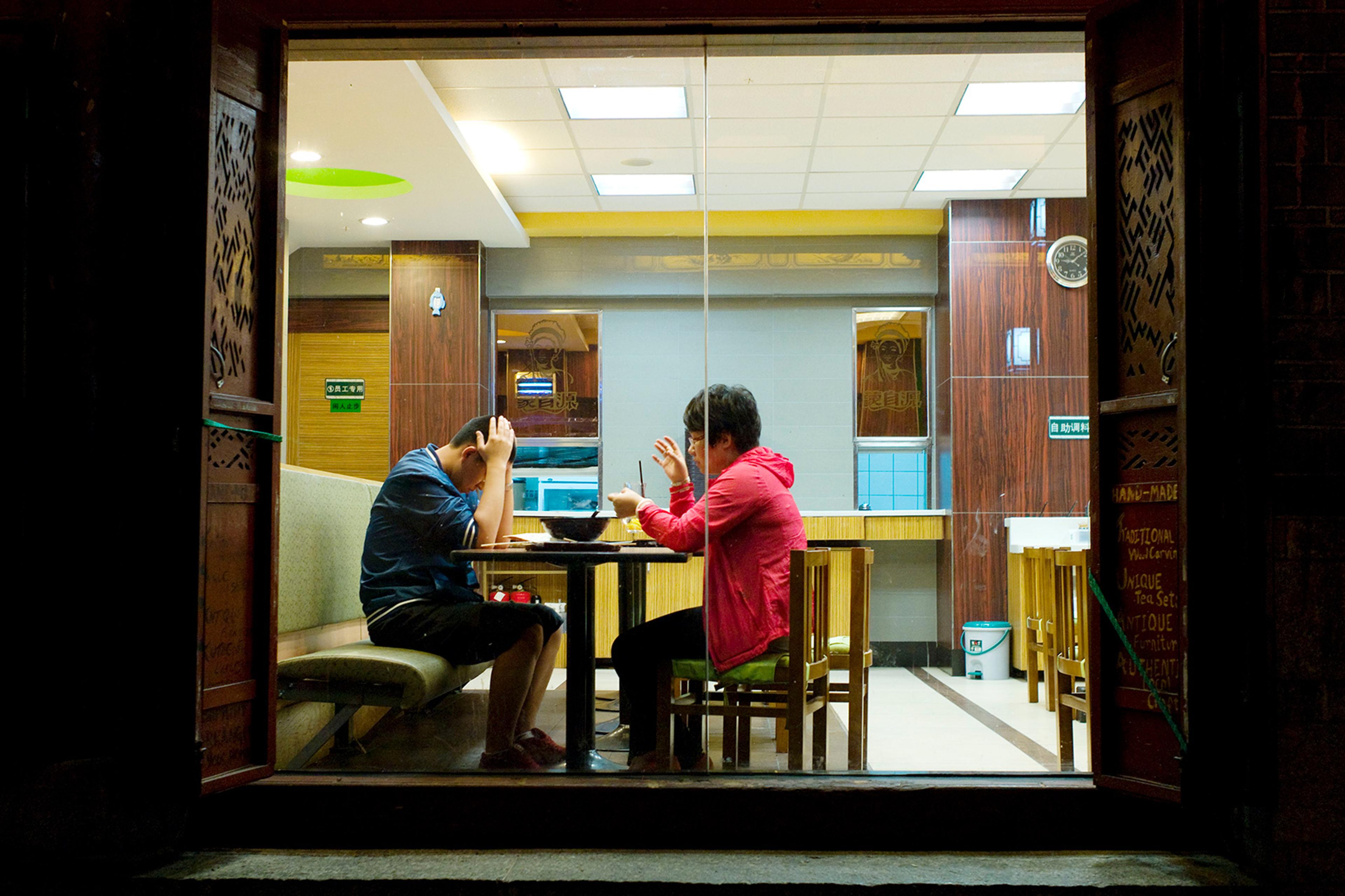 Two people facing each other in a restaurant, one gesticulates, the other has head in his hands, seen through a window at night.