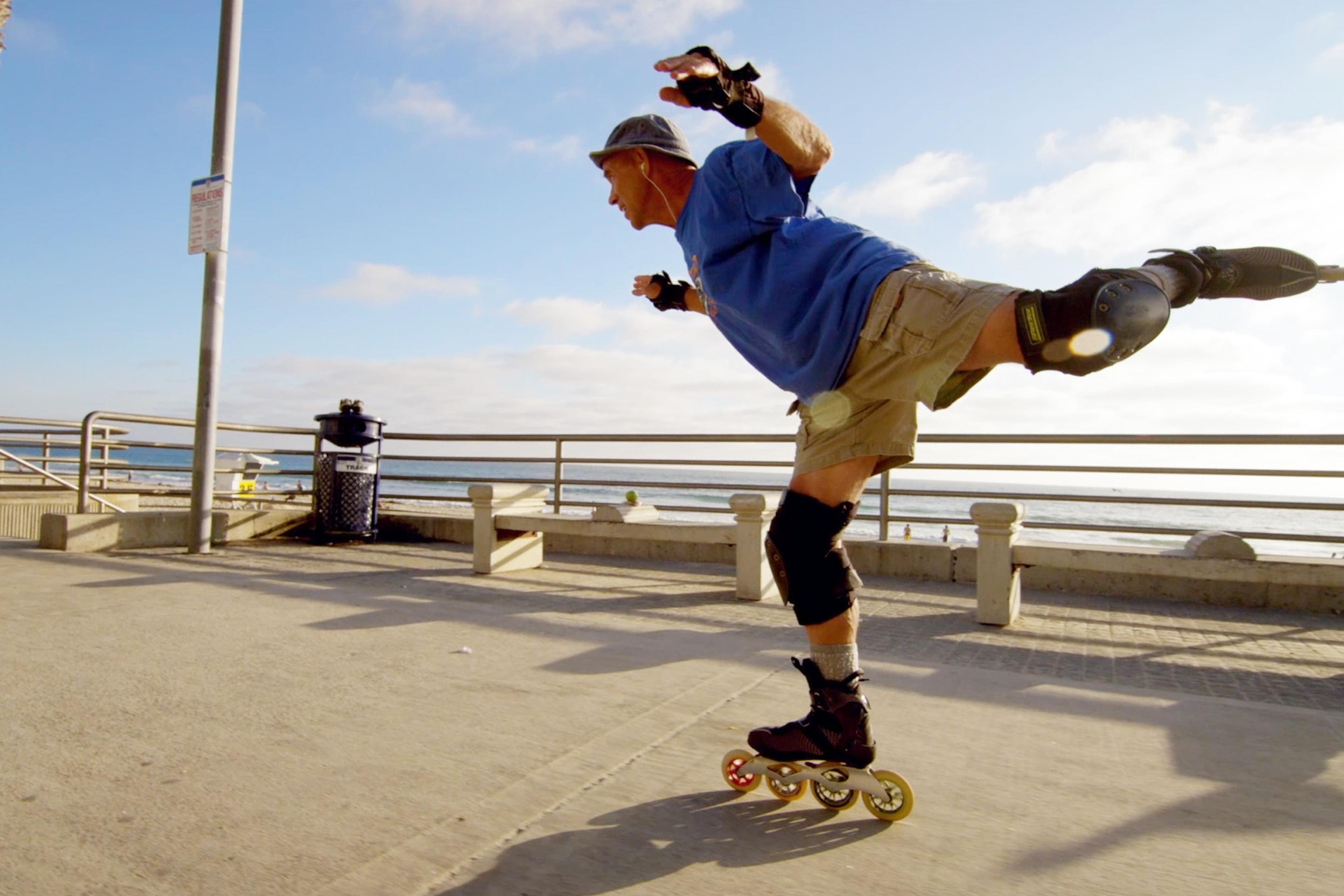Person roller-skating by the seaside, wearing protective gear and balancing on one foot, with a balustrade, palm tree, and sunny sky in the background.