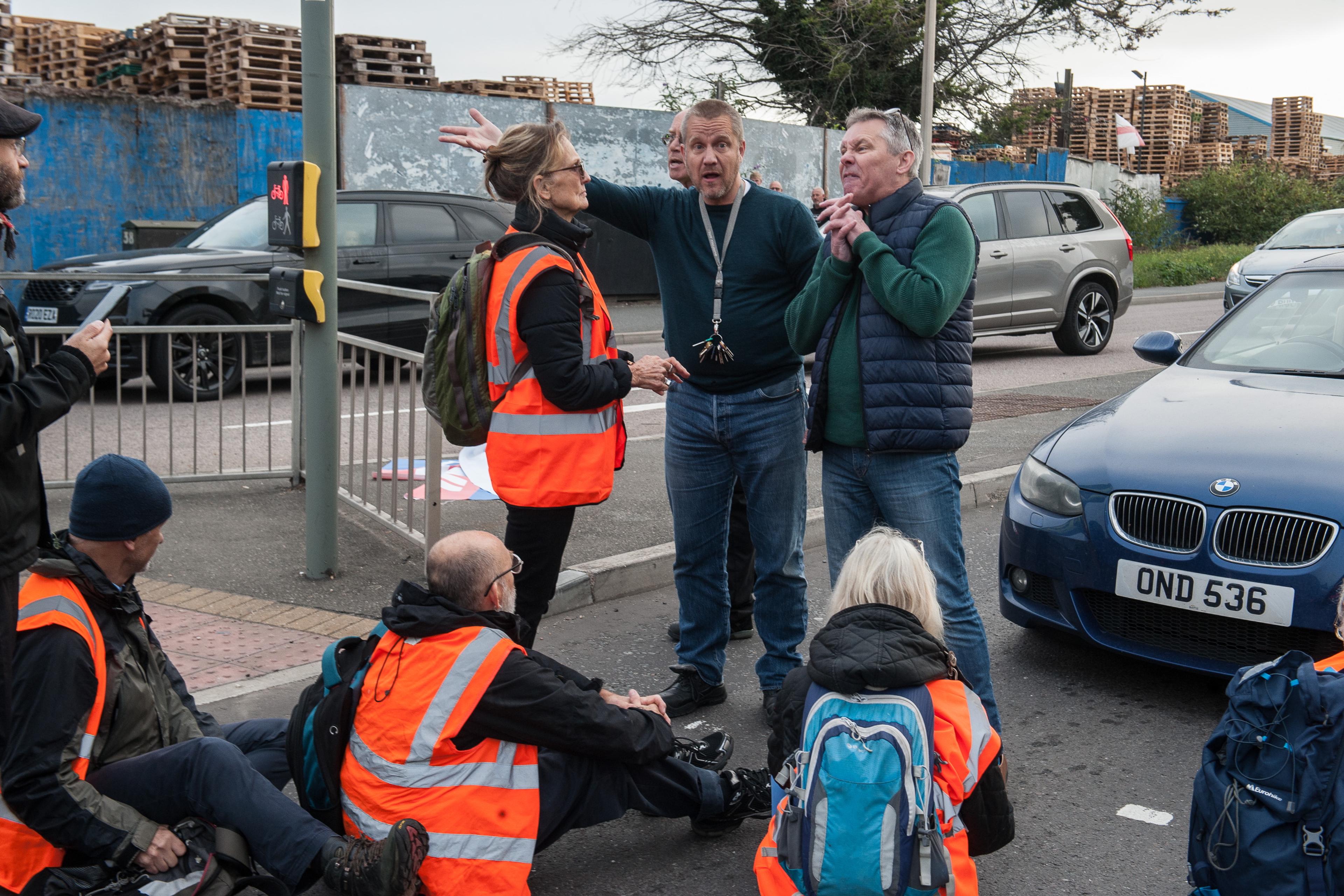 Photo of people in high-vis vests sitting on a road, engaged in discussion, with cars and wooden pallets in the background.