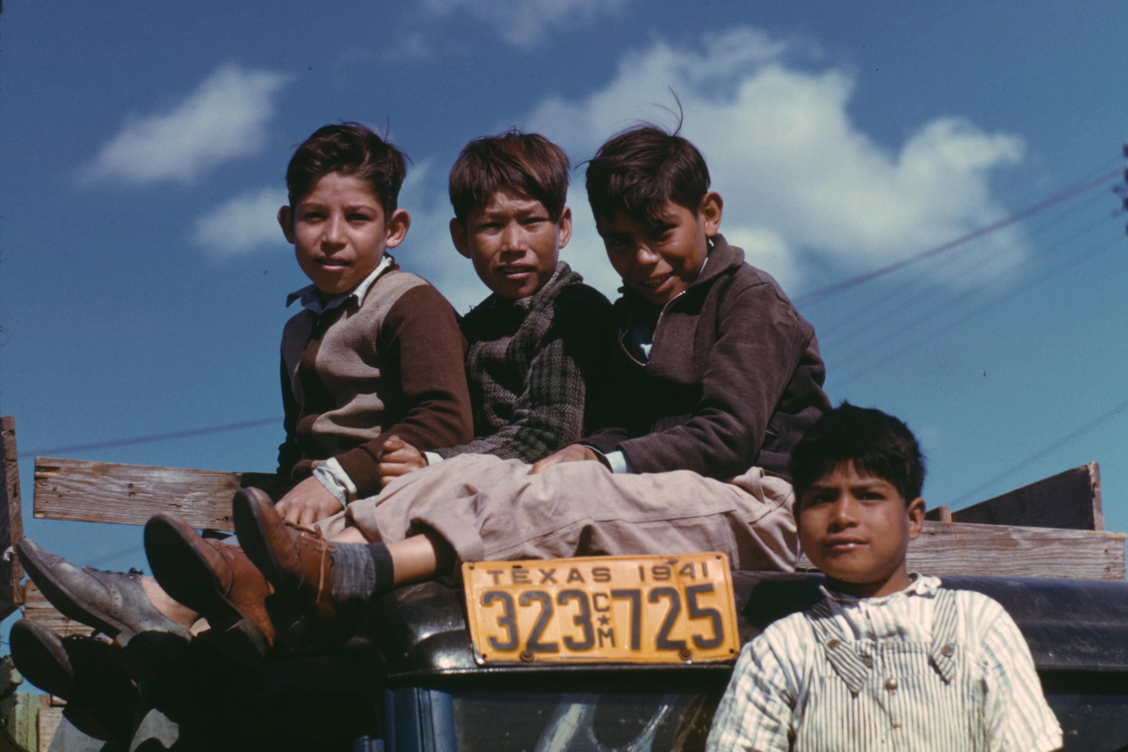 Photo of four boys sitting on a wooden board of a vehicle with a 1941 Texas number plate against a blue sky.