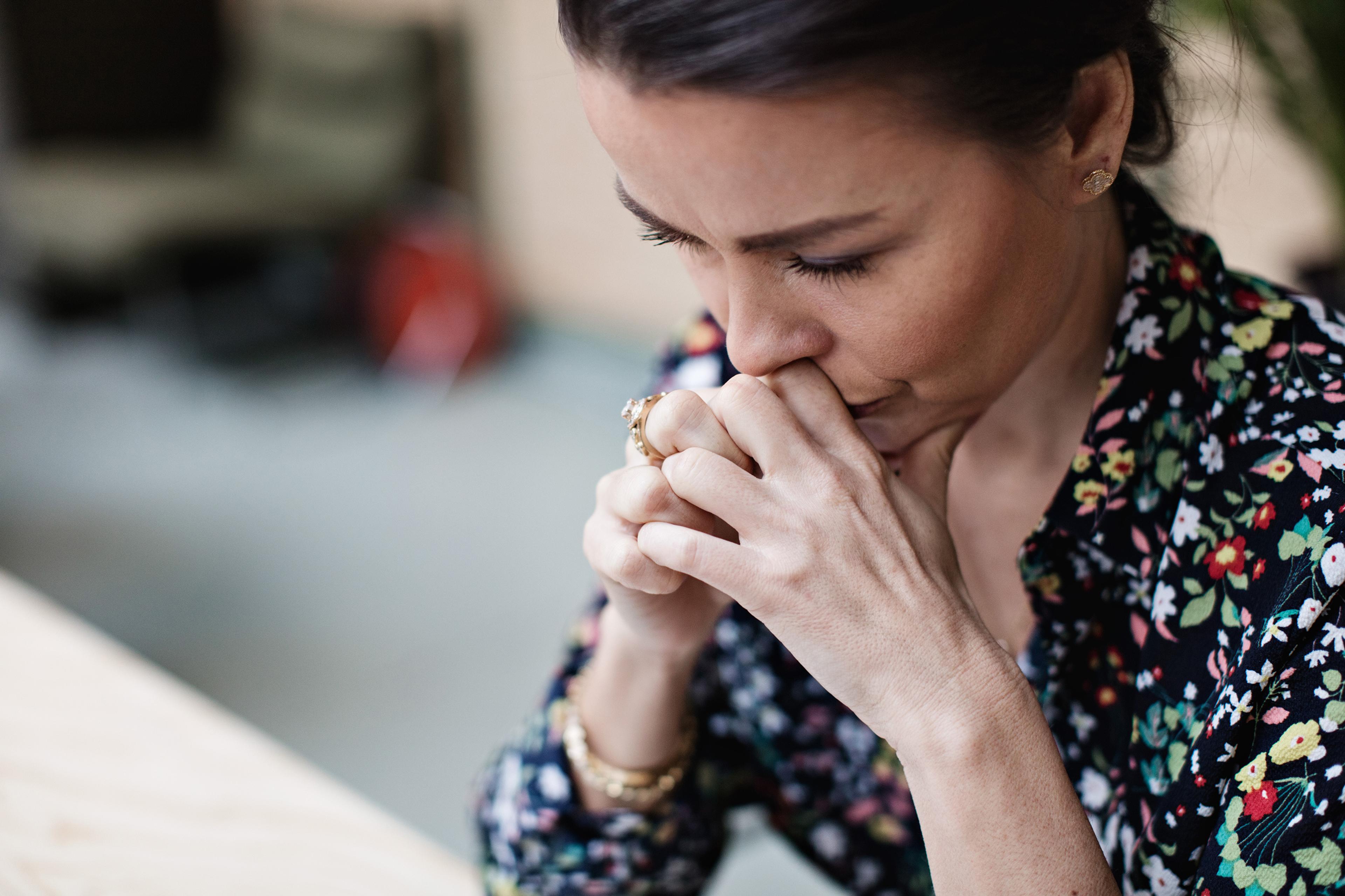Photo of a woman in a floral blouse thinking deeply with her hands clasped near her face.