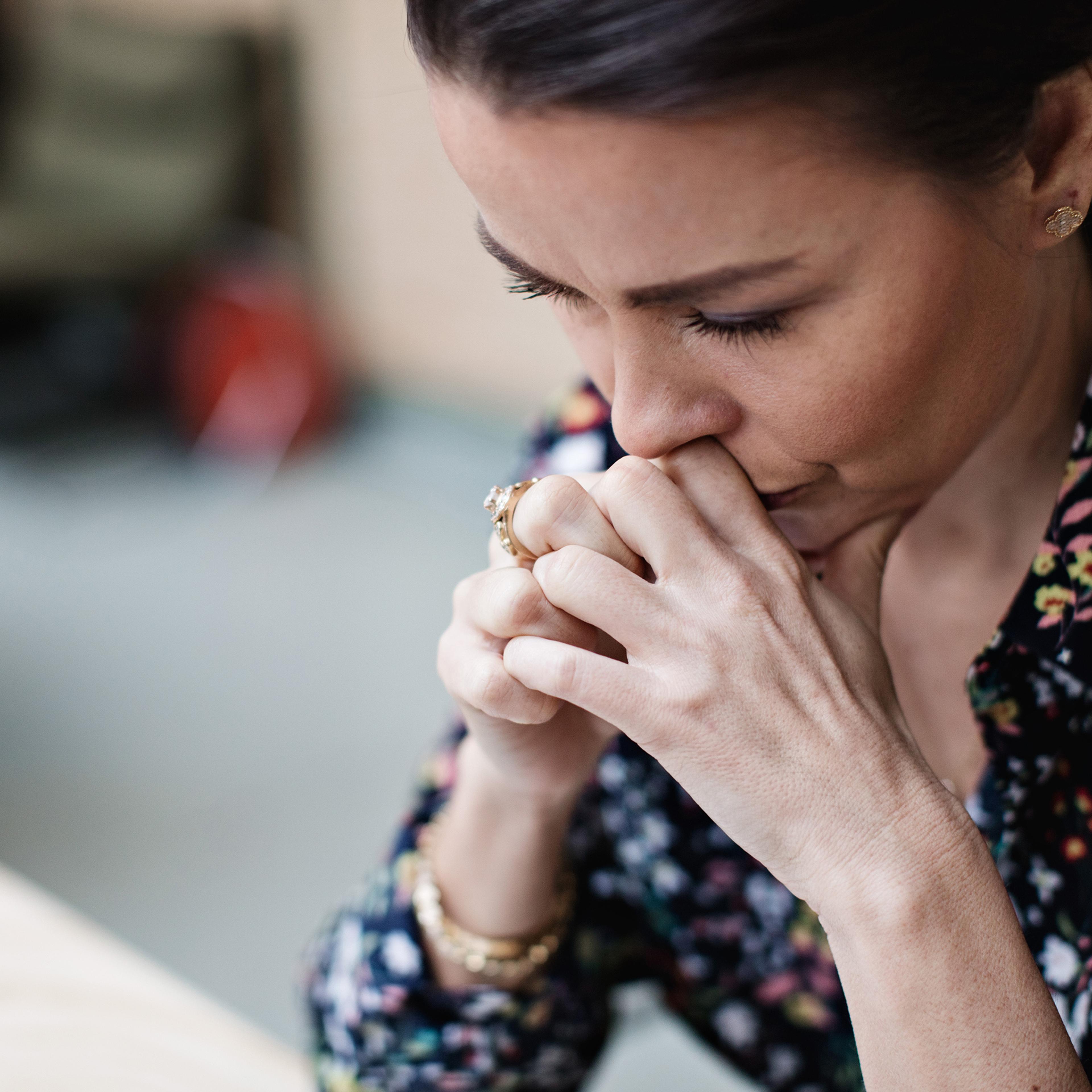 Фотография женщины в цветочной блузке, глубоко задумавшейся, сложив руки у лица.