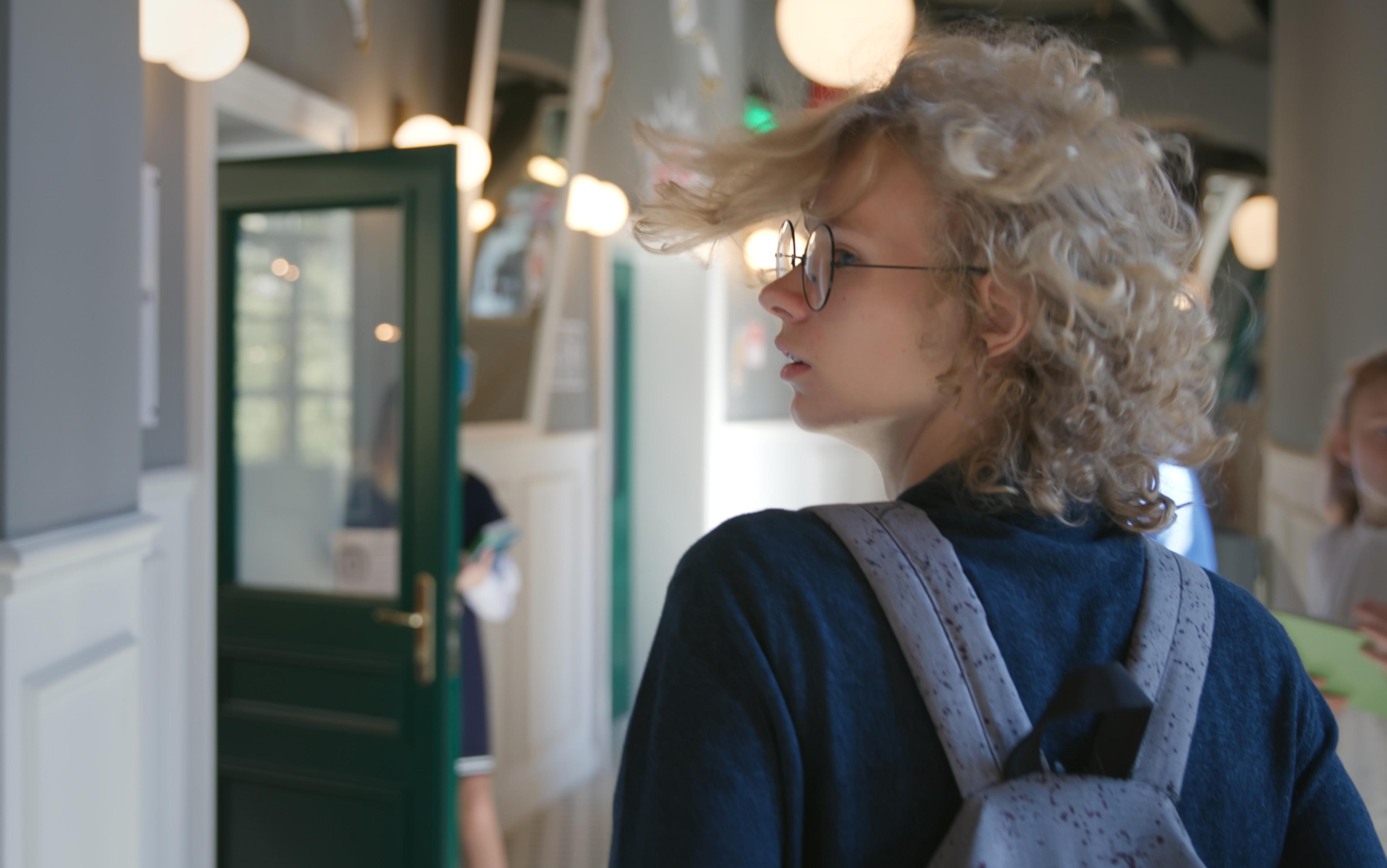 A young person with curly hair and glasses, wearing a backpack, indoors in what appears to be a school setting, with a green door and soft lighting, other children in background.