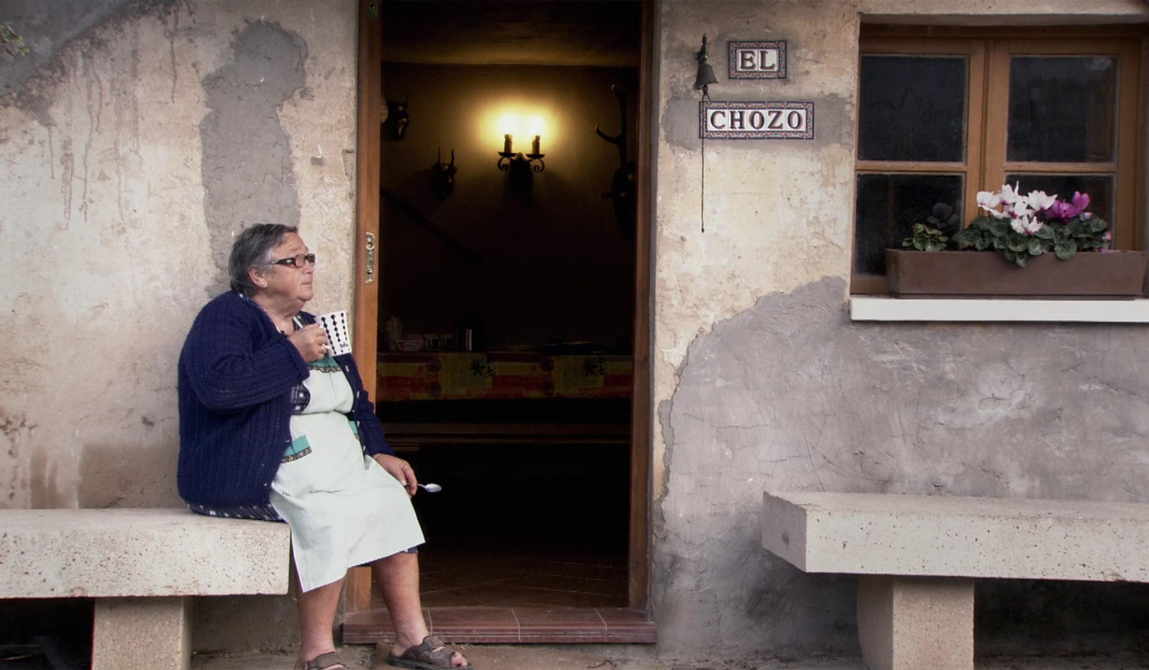An elderly woman in a blue cardigan and apron sits outside a rustic building with a mug, sign, and window box flowers.