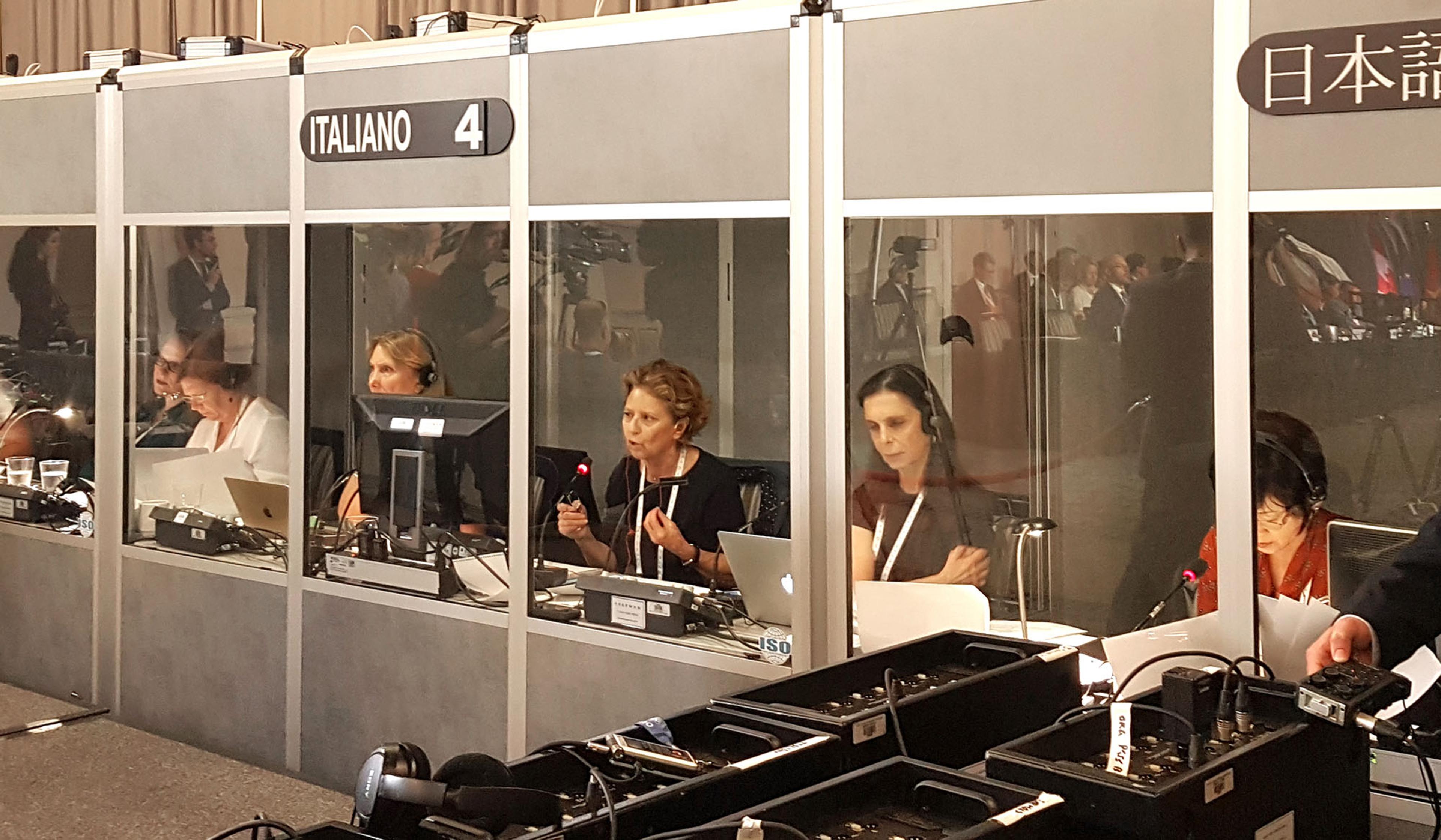 Interpreters working in separate booths for different languages, equipped with computers, microphones, and headsets at a conference.