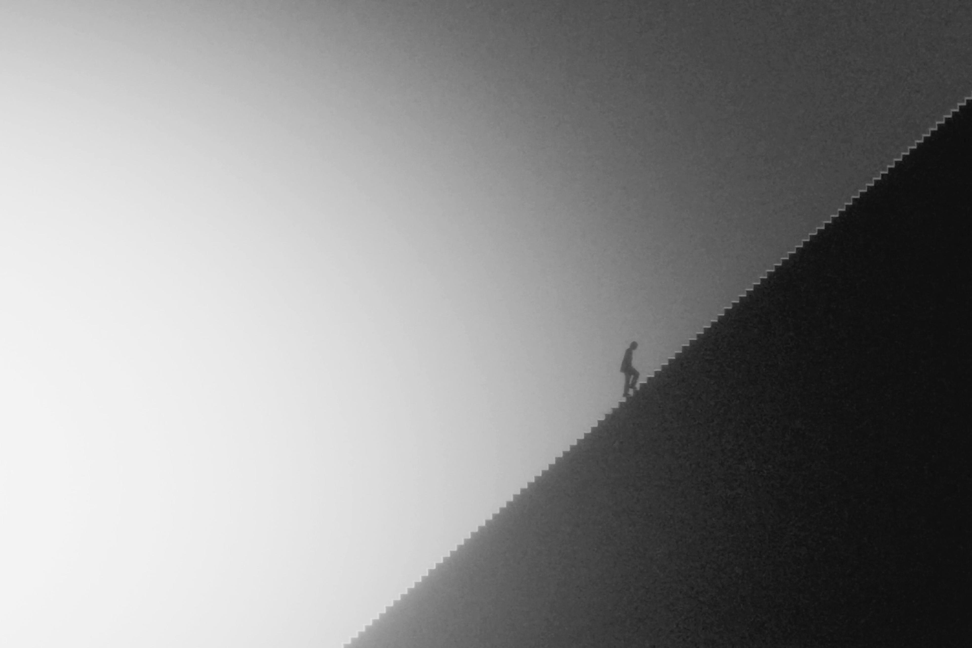 Silhouette of a person climbing a steep, diagonal staircase against a gradient sky in grayscale.