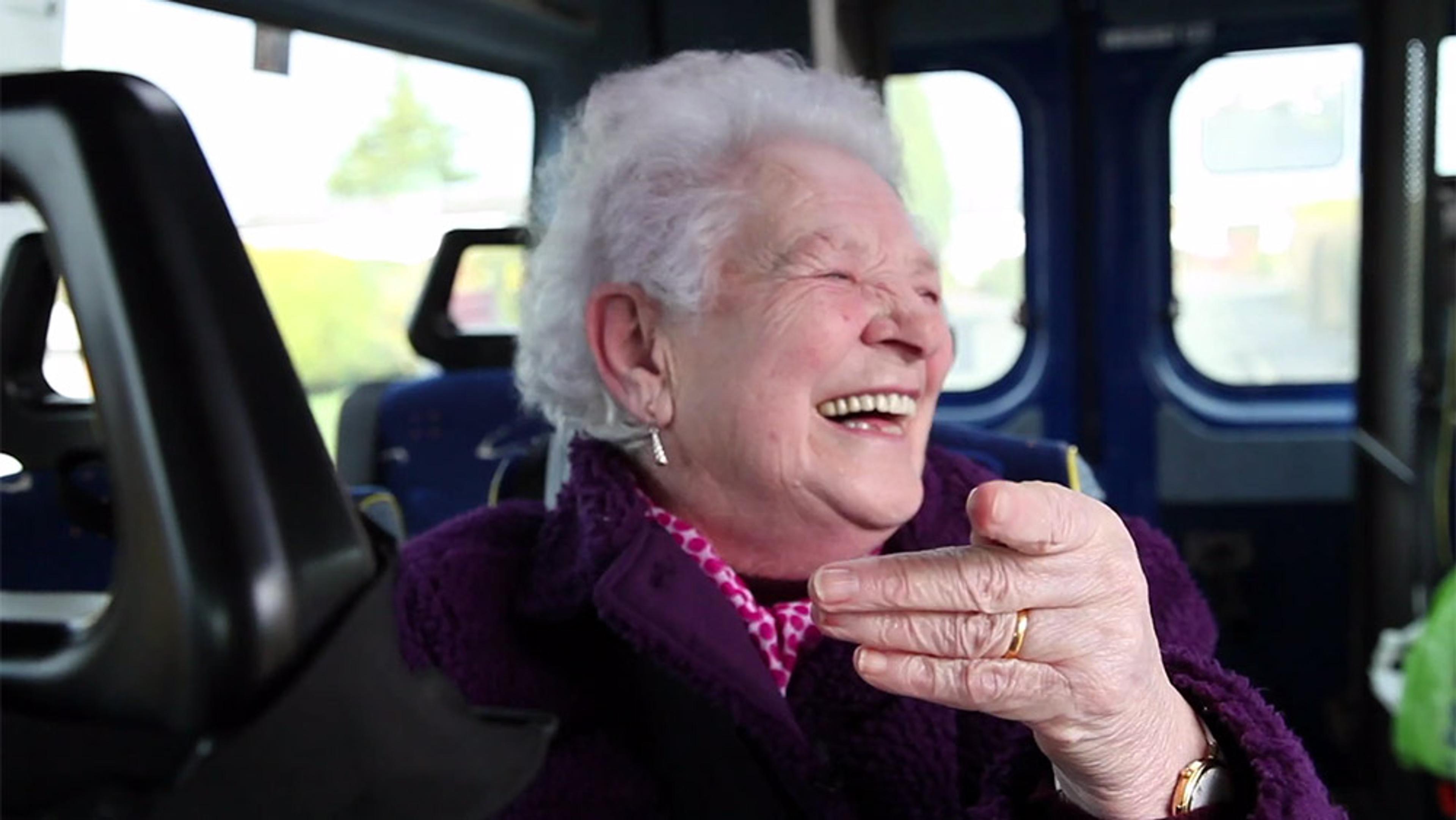 A cheerful elderly woman with short white hair, laughing on a bus, wearing a purple coat and pink scarf.