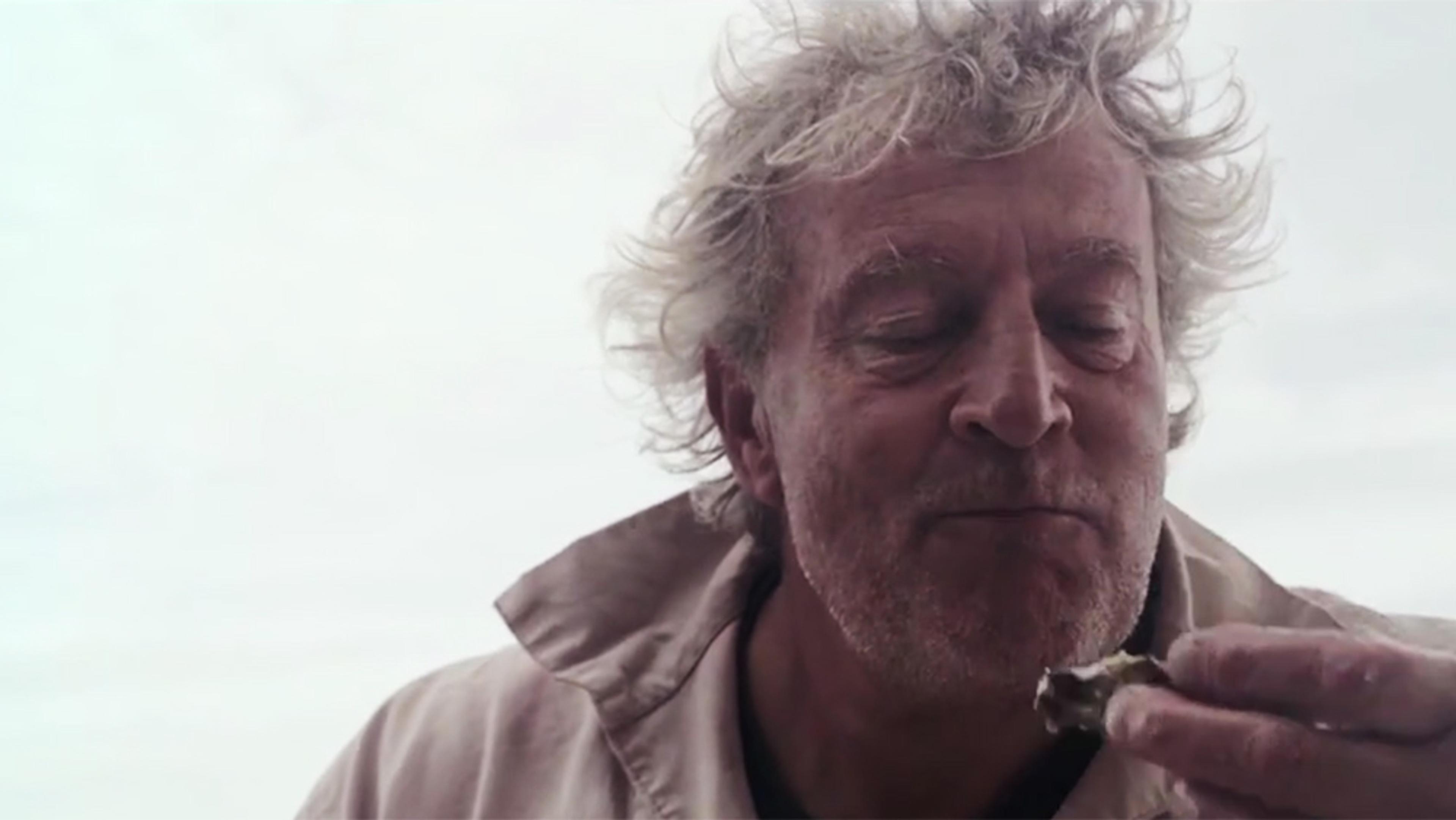 An elderly man with tousled grey hair eating an oyster, wearing a beige jacket, eyes closed in apparent enjoyment.