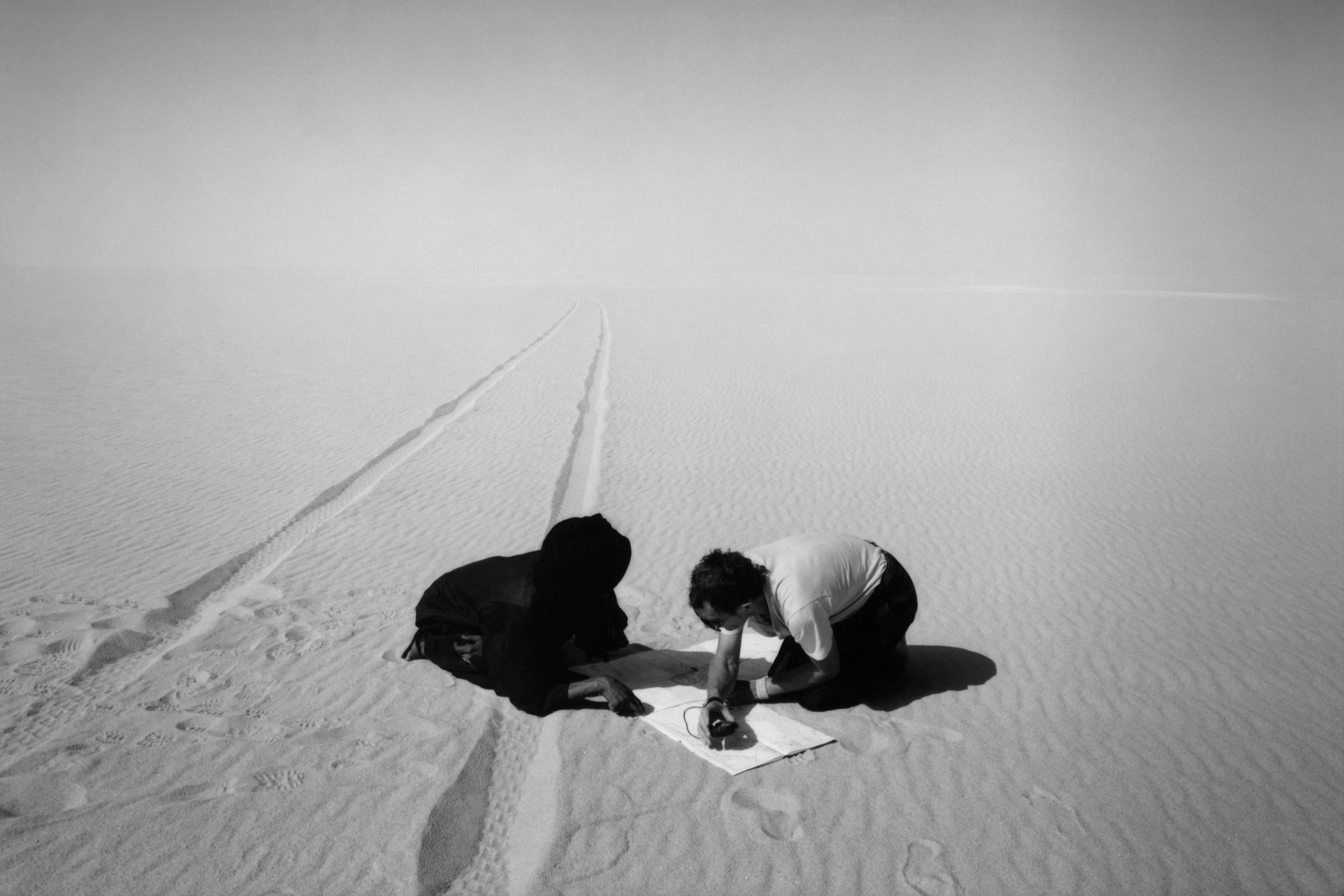 Black-and-white photo of two people kneeling on sand examining a map with visible tracks in a vast desert landscape.