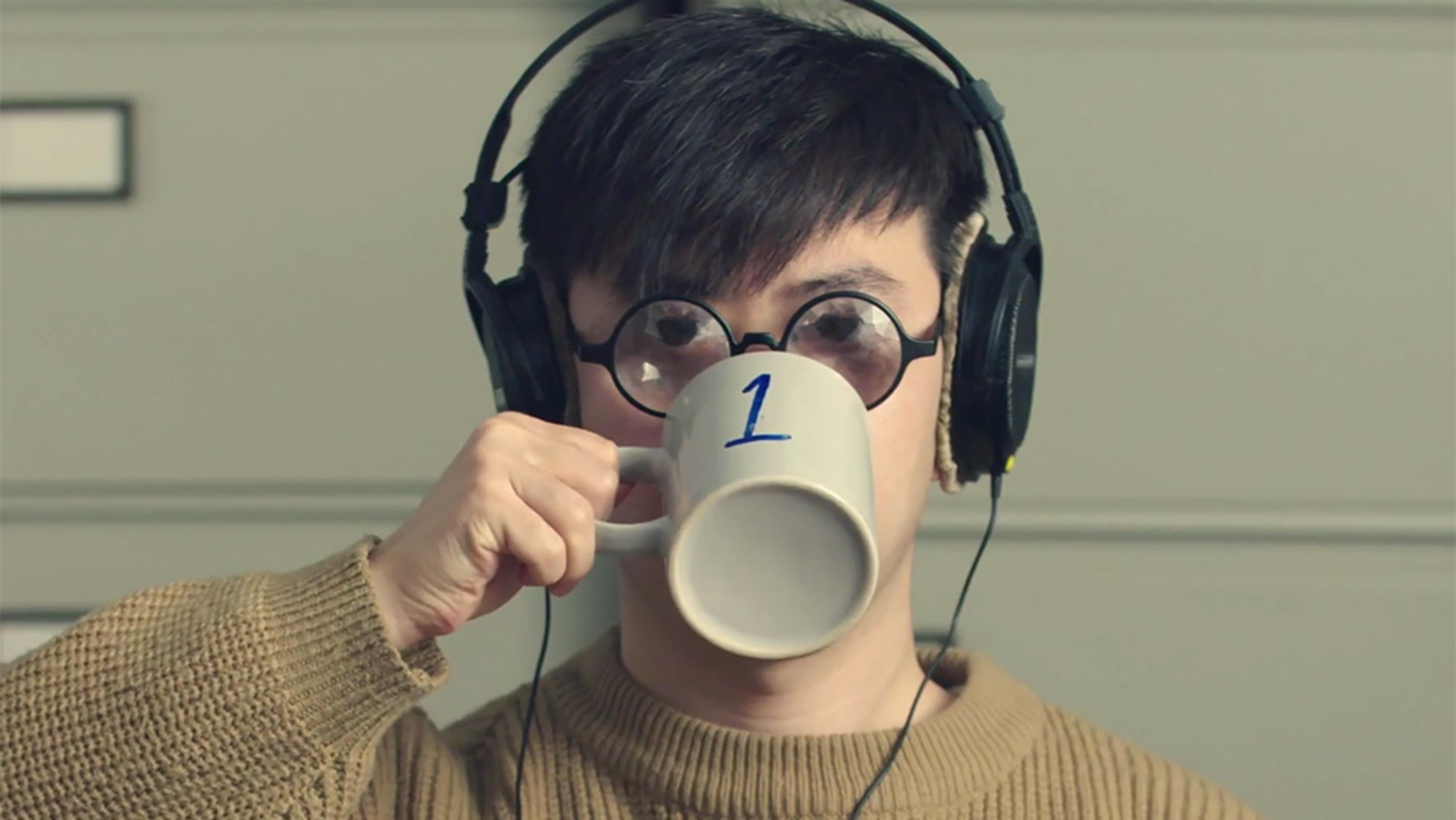 A person wearing glasses and headphones, drinking from a mug with the number 1 on it. They wear a brown jumper.