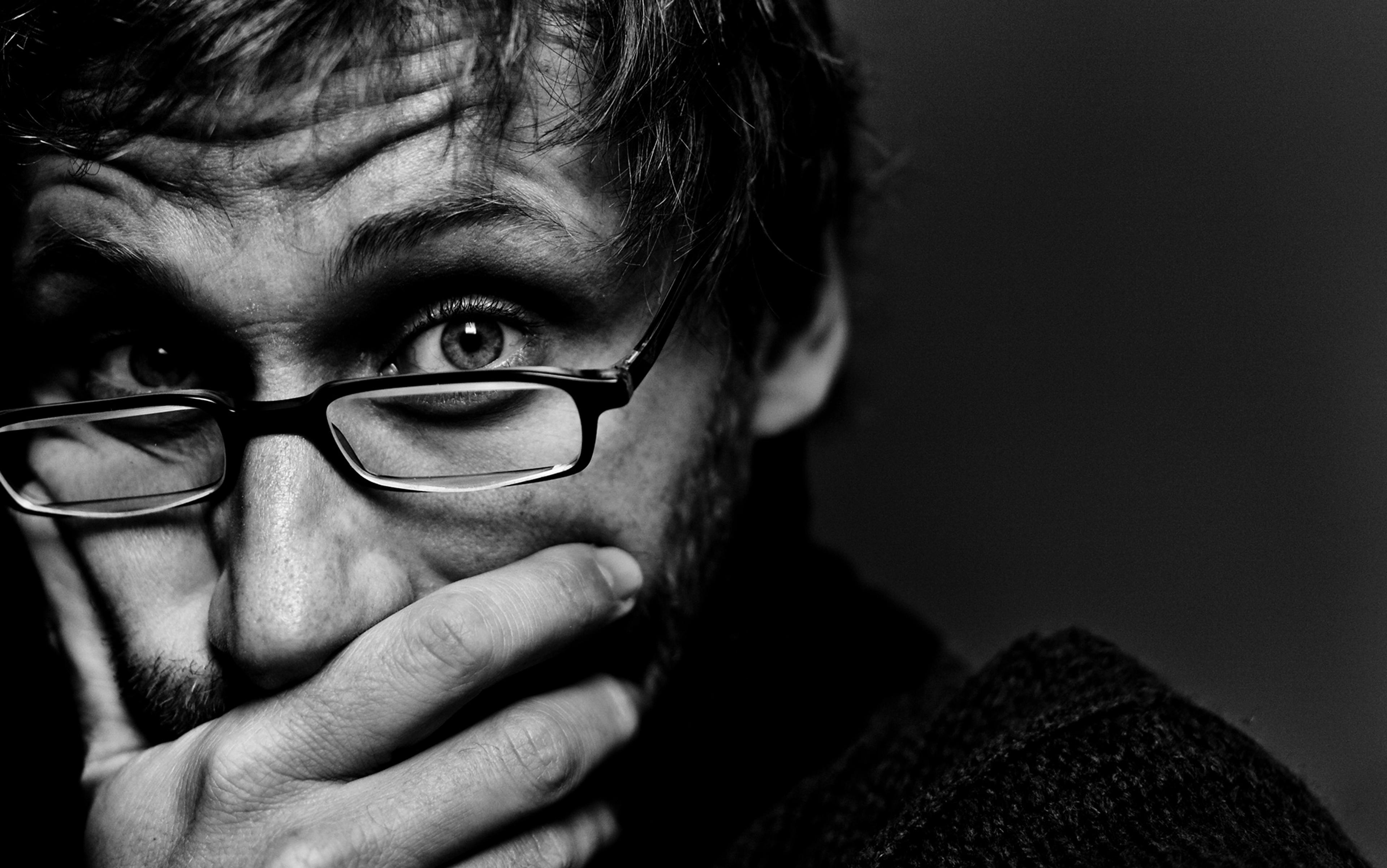 Black and white close-up of a person with glasses, covering mouth with their hand, looking intensely at the camera.