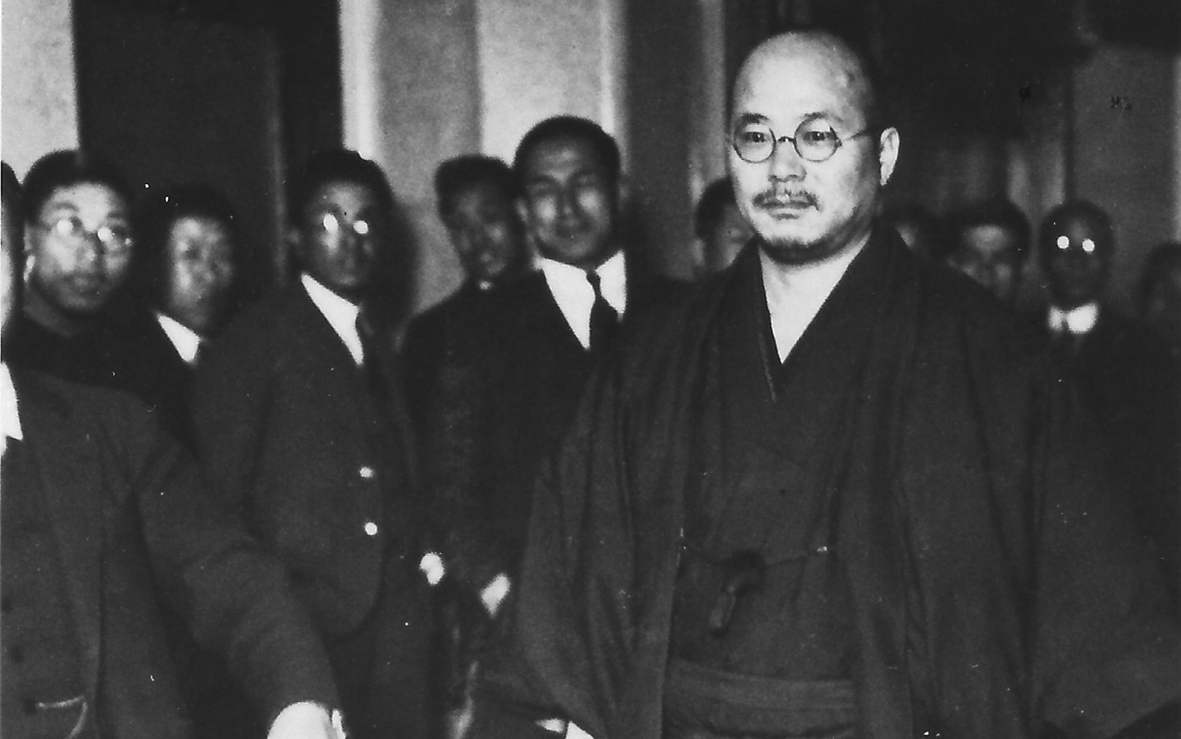 Black and white photo of a bald Japanese man in traditional attire, wearing glasses, surrounded by a group of men in suits and ties.