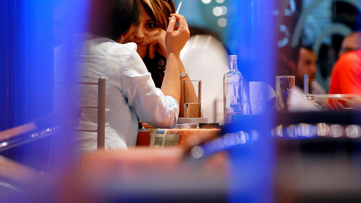 Two people at a cafe table, one holding a cigarette. Blurred foreground and background create a candid atmosphere.