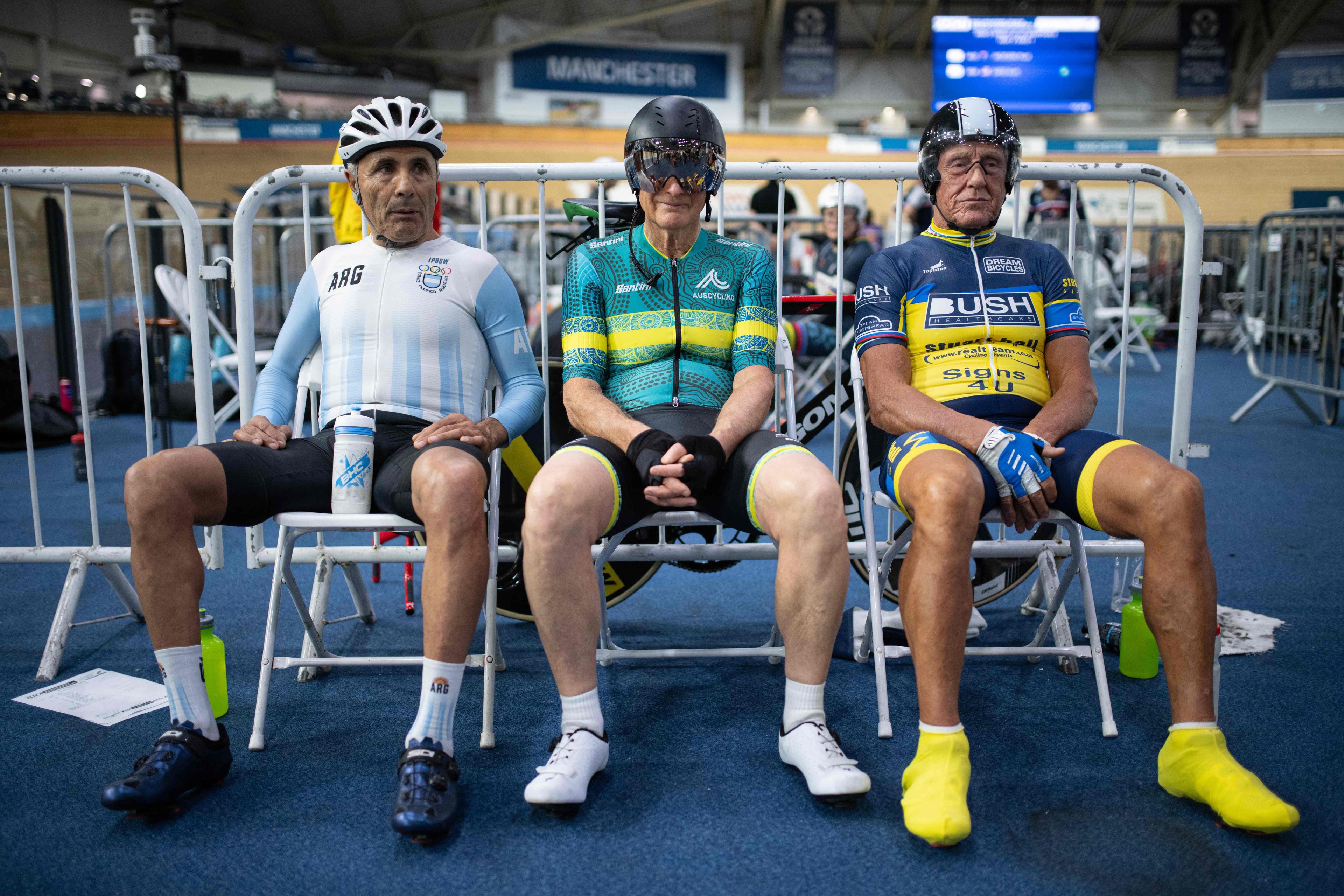 Photo of three elderly cyclists in racing gear seated indoors at a velodrome in Manchester.