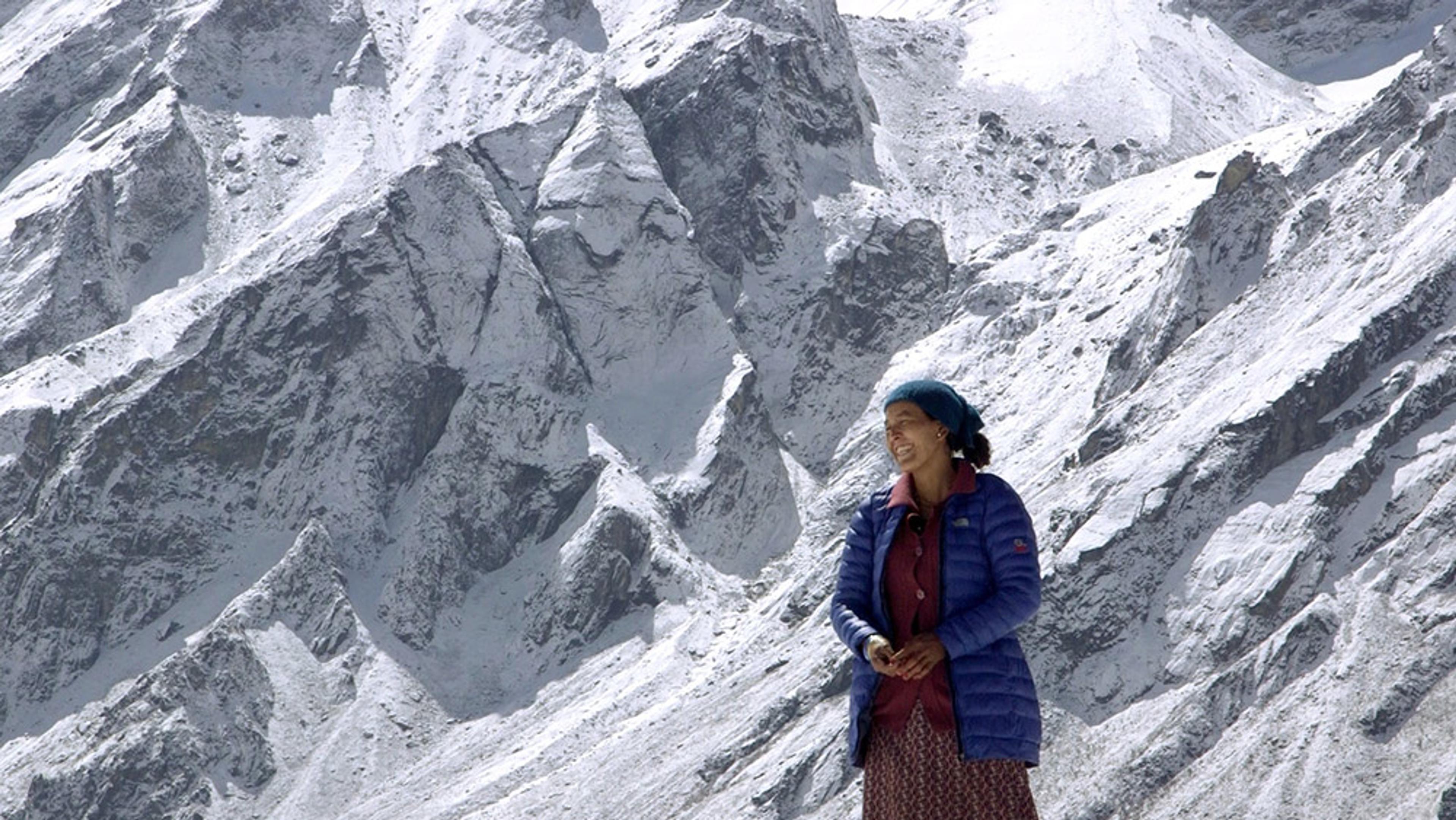 A woman standing in front of towering, snow-covered mountains, wearing a blue jacket and a blue hat, smiling.