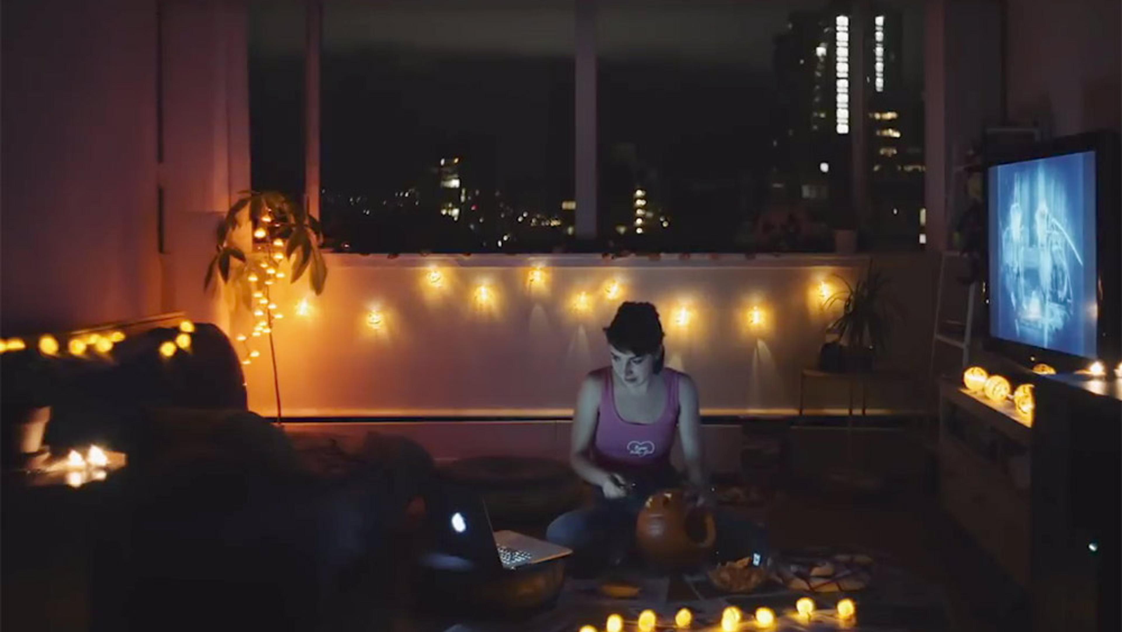 A young woman carving a pumpkin at night in a cosy, dimly lit room with fairy lights, a laptop and a TV displaying a black and white film.