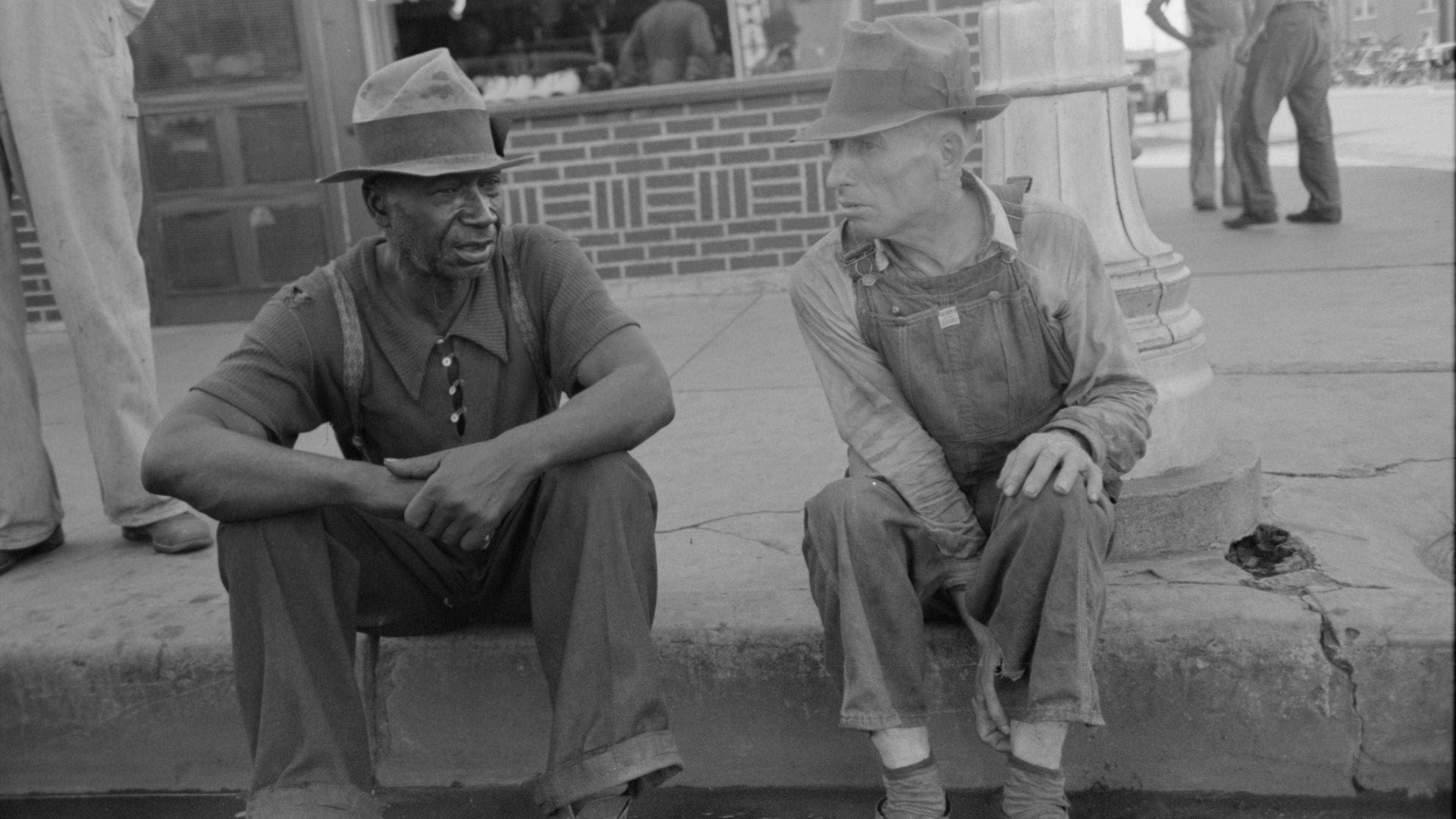 Black and white photo of two men sitting on a kerb in conversation with a brick storefront behind them.