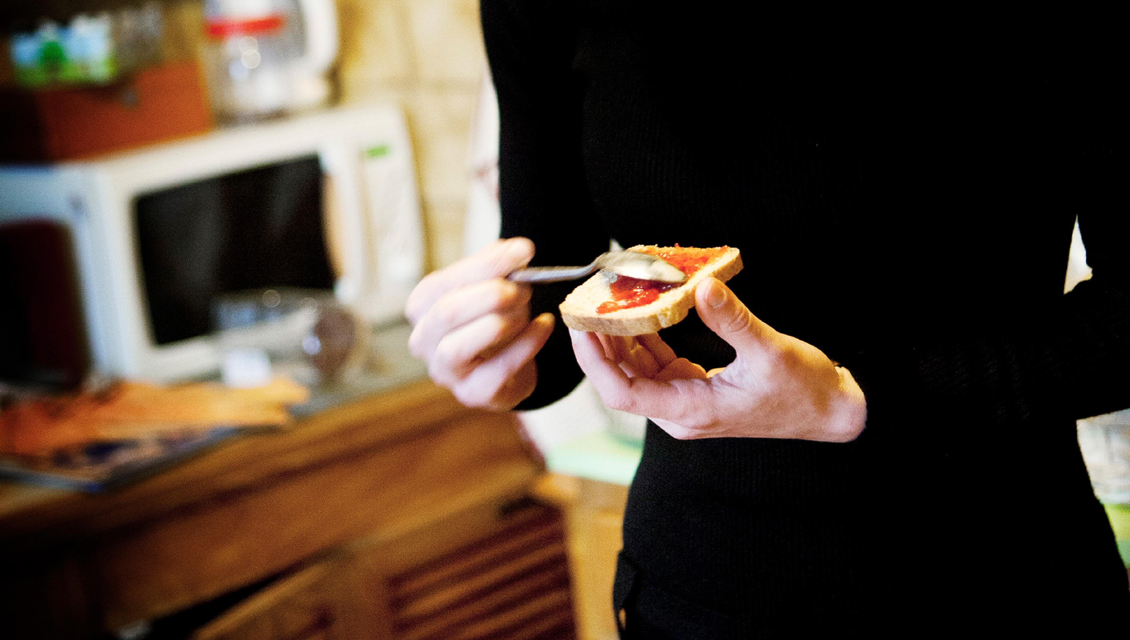 Person spreading strawberry jam on a piece of toast with a spoon in a kitchen setting, with a microwave and wooden countertop in the background.