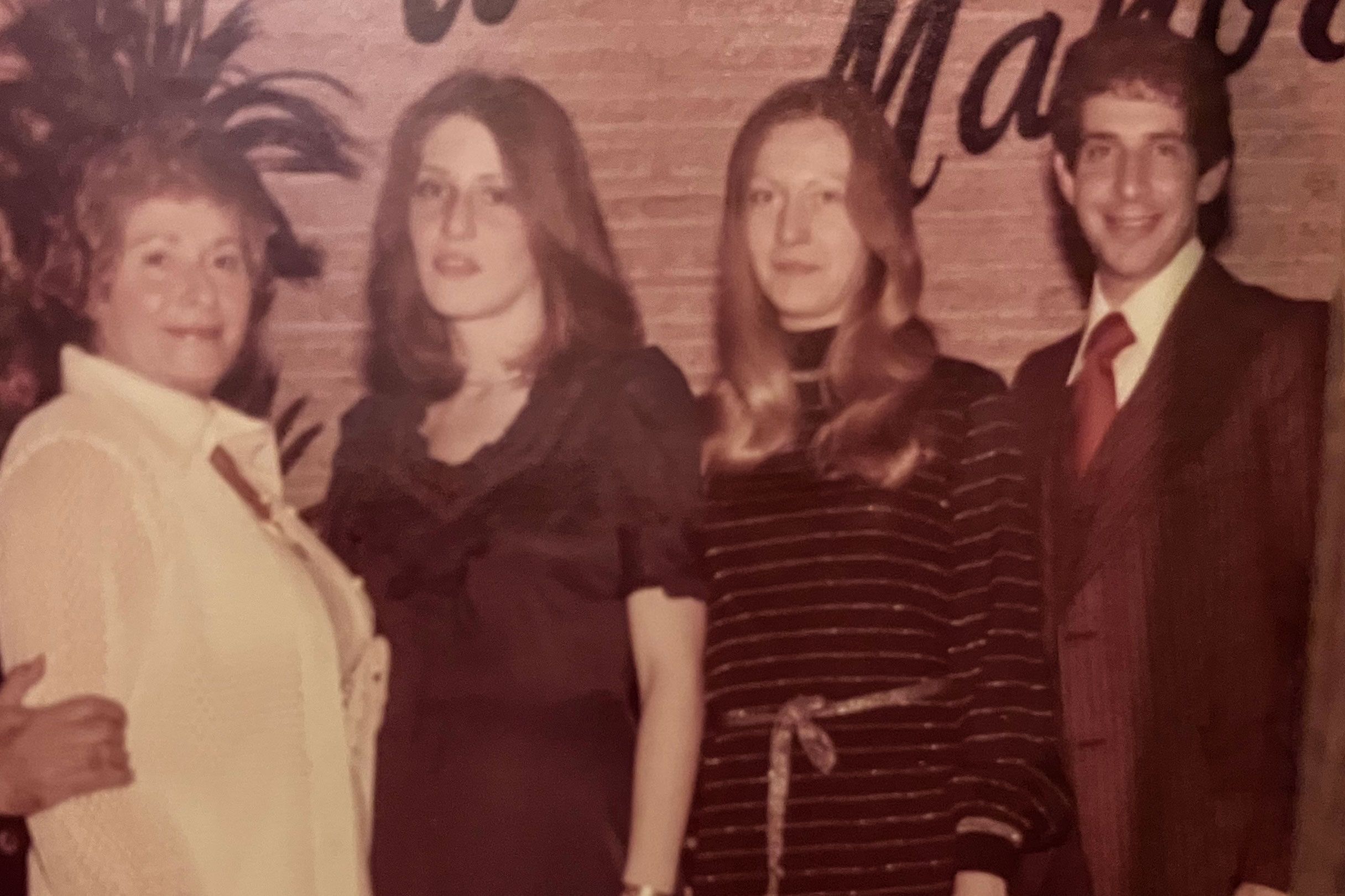 Vintage photo of four people posing indoors with a plant and brick wall background.