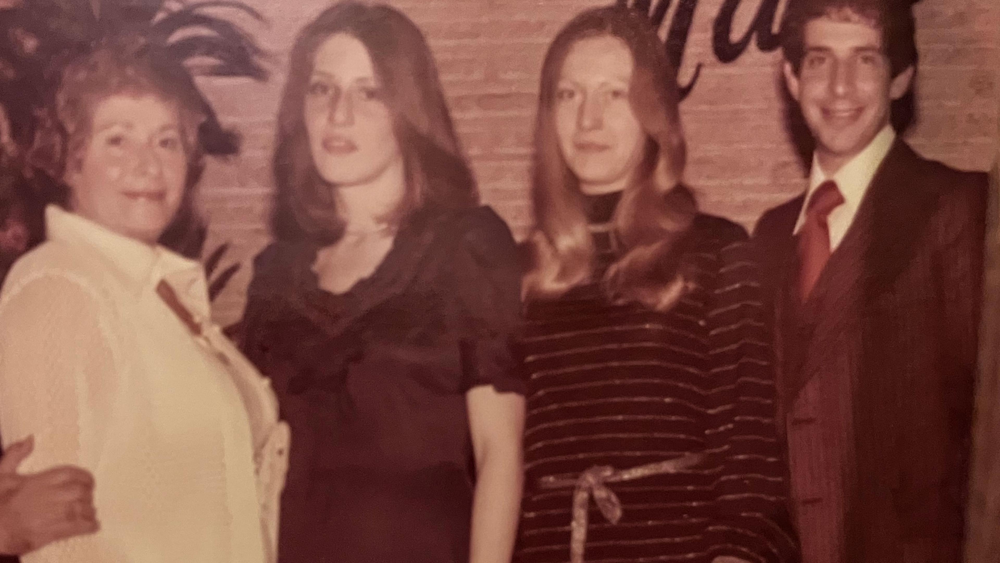 Vintage photo of four people posing indoors with a plant and brick wall background.