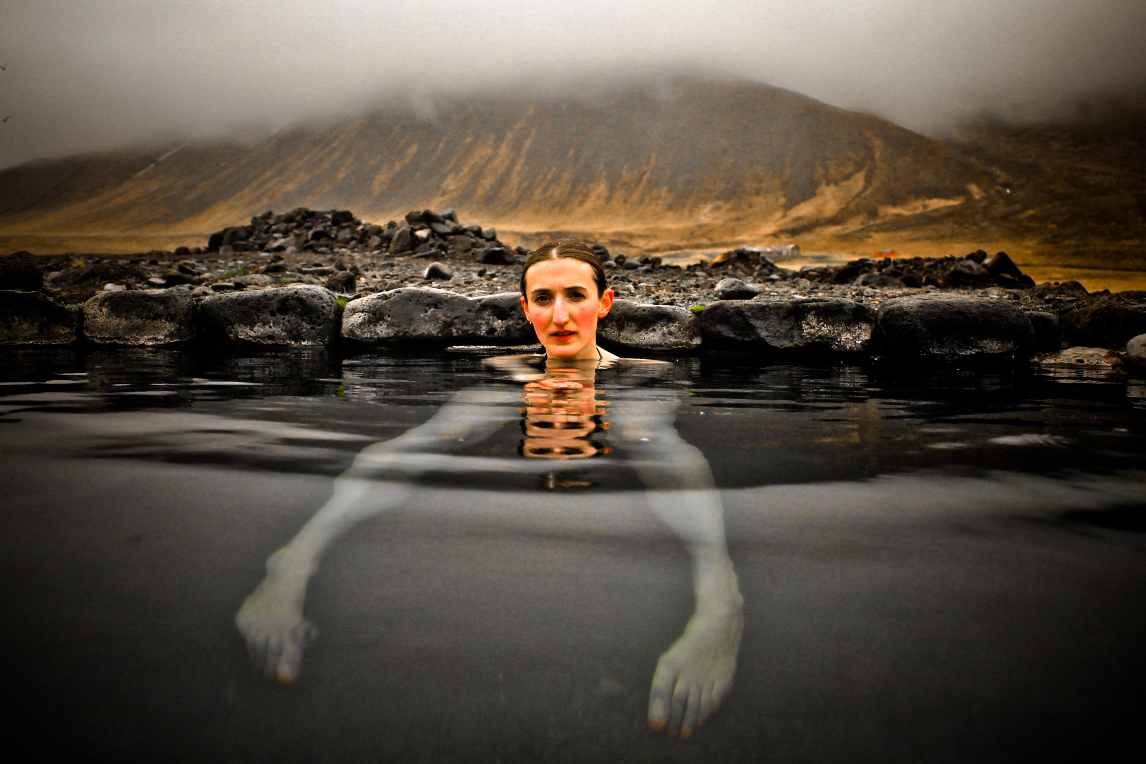Photo of a woman submerged to the neck in a natural hot spring, with mountains and mist in the background, creating a serene atmospheric scene.