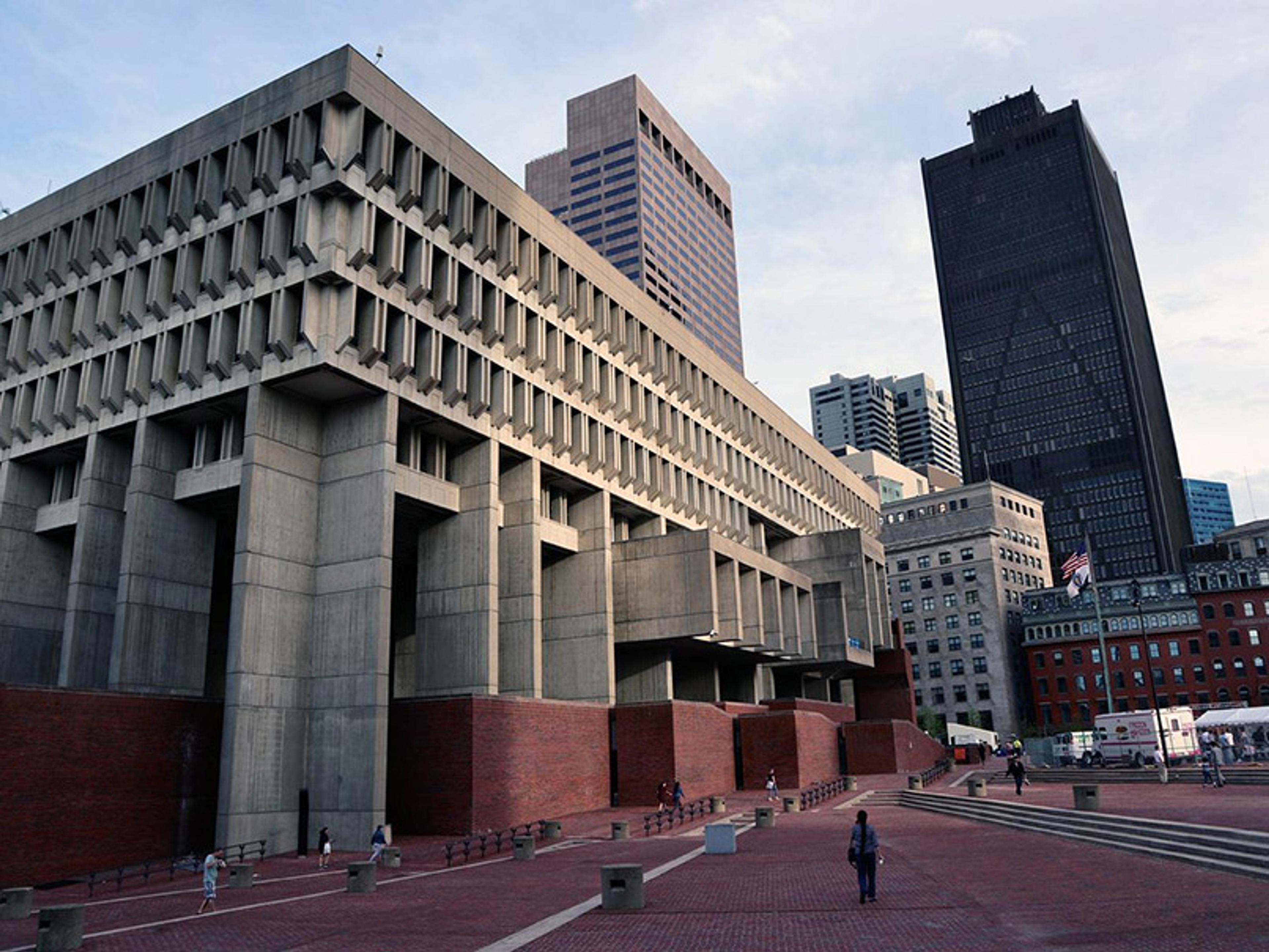 A Brutalist concrete building with an angular design, a large plaza in front, and taller modern buildings in the background.