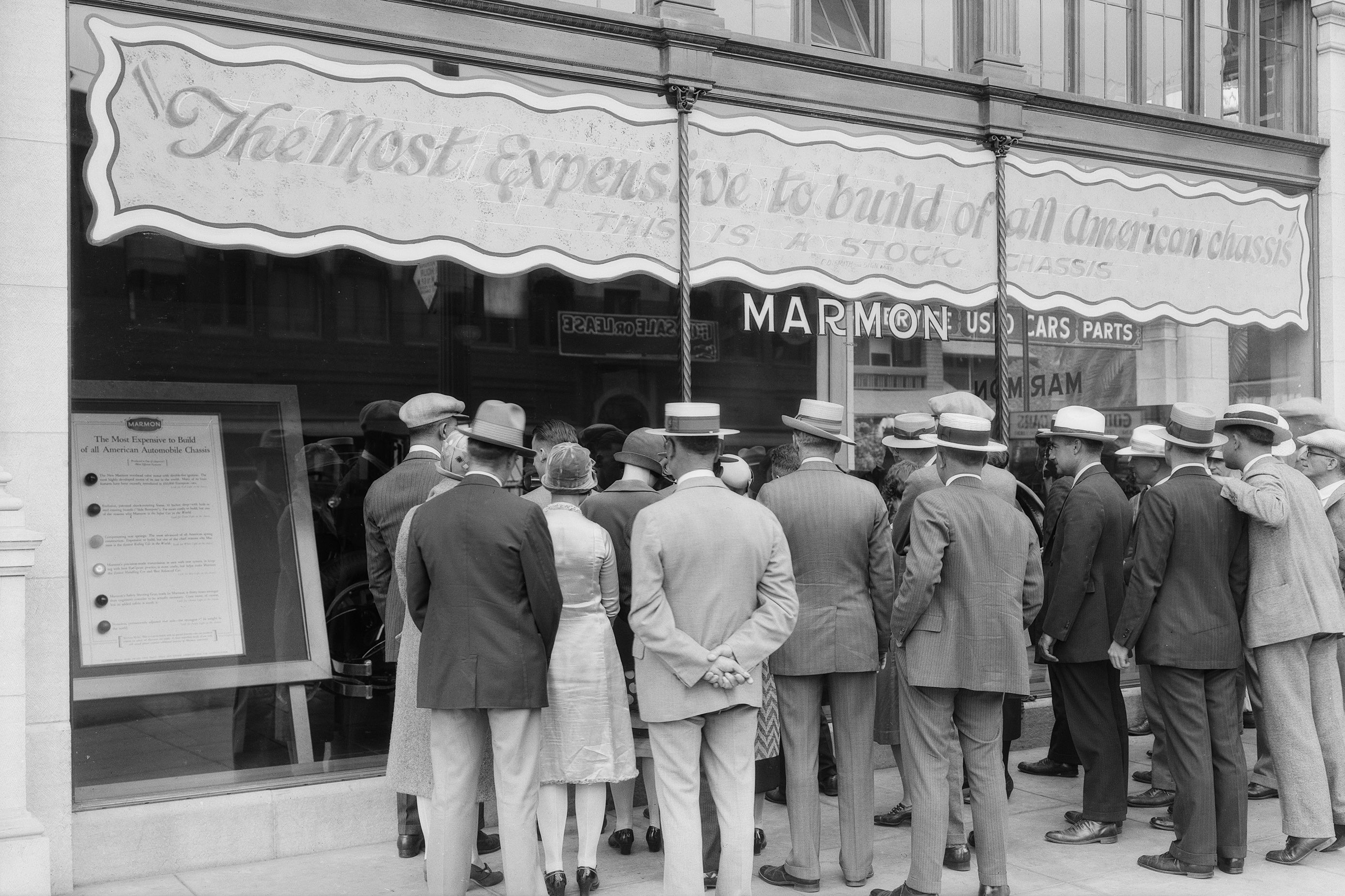 Black and white photo of a crowd gathered outside a Marmon car showroom, reading a display about expensive car chassis.