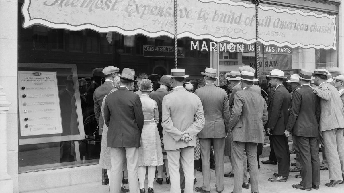 Black and white photo of a crowd gathered outside a Marmon car showroom, reading a display about expensive car chassis.