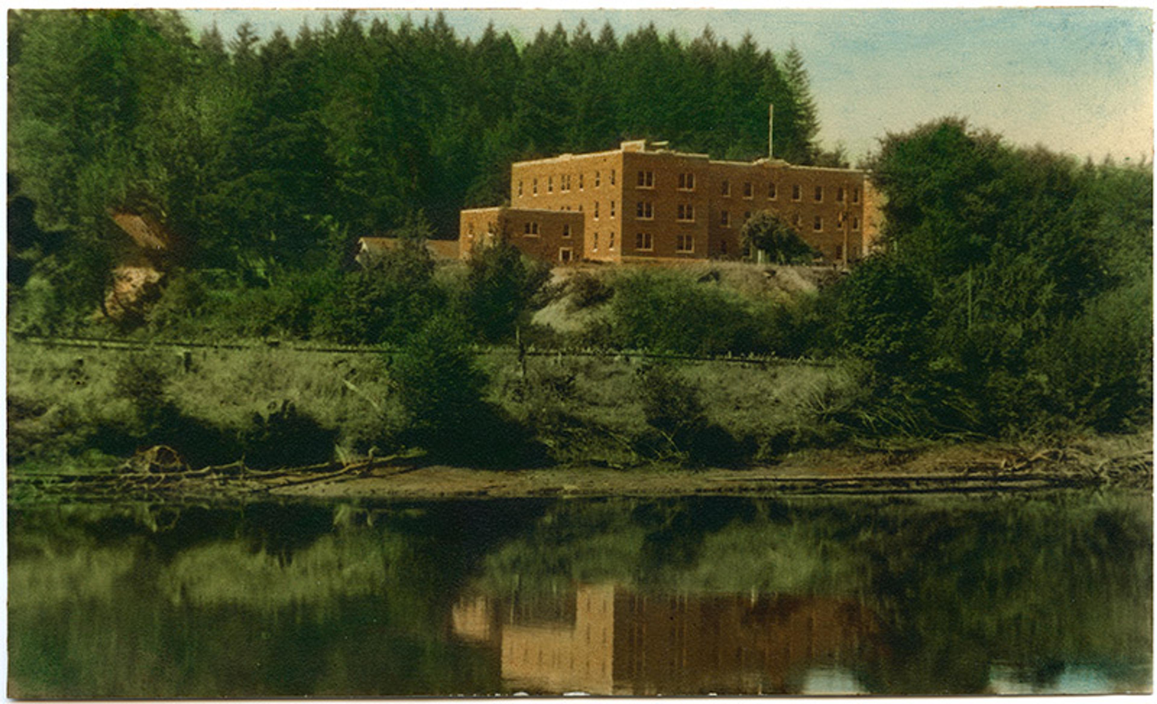Painting of a brick building surrounded by trees reflected in a calm lake with a wooded area in the background.