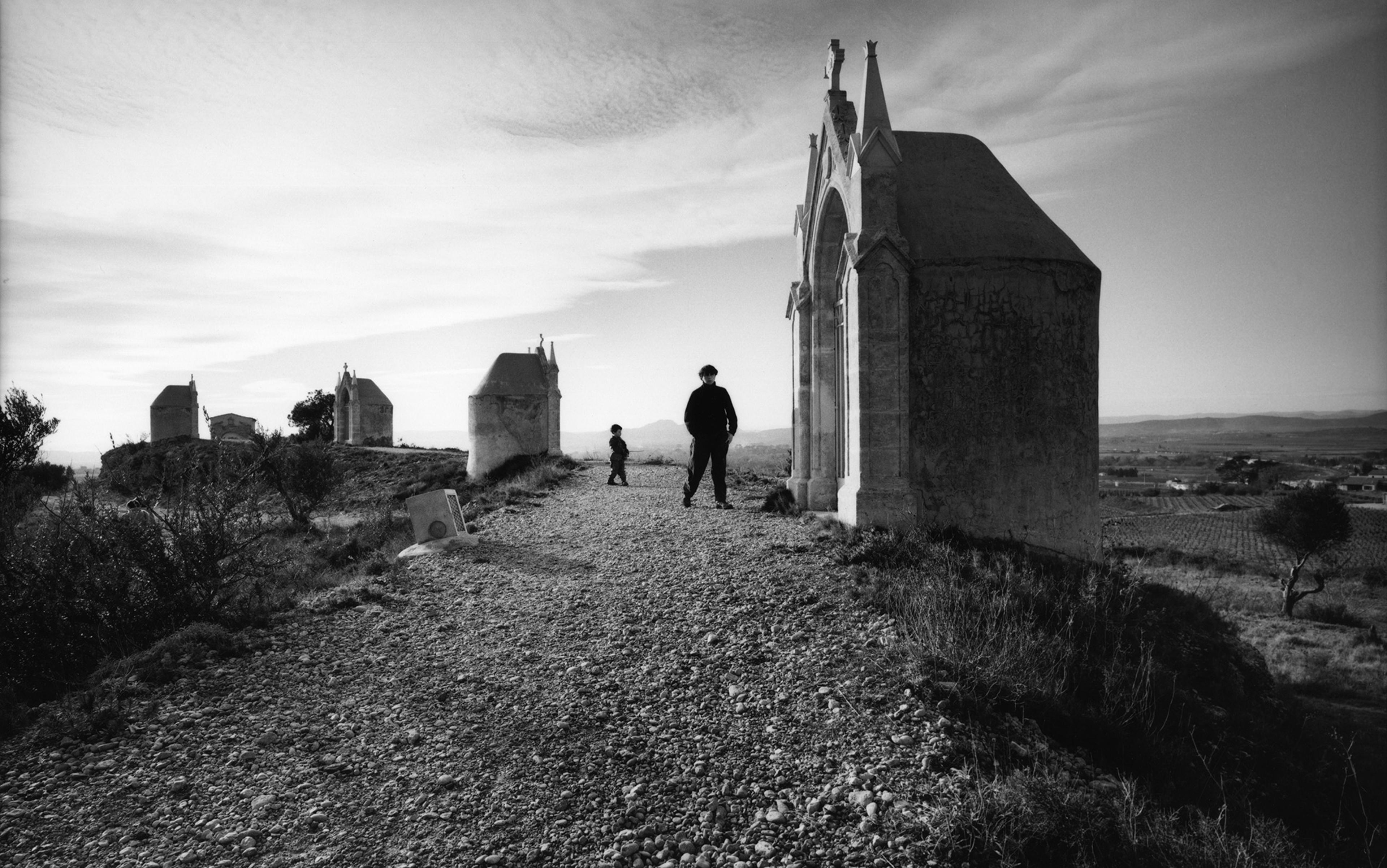Black-and-white photo of adult and child walking among old stone structures on a rocky path, set against an expansive landscape.