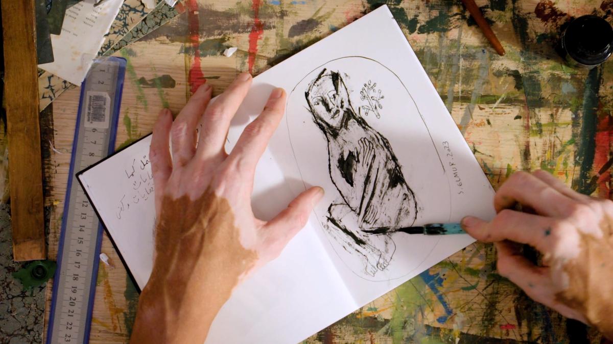 Photo of hands drawing a figure in a sketchbook on a splattered art table with ruler and paintbrush.