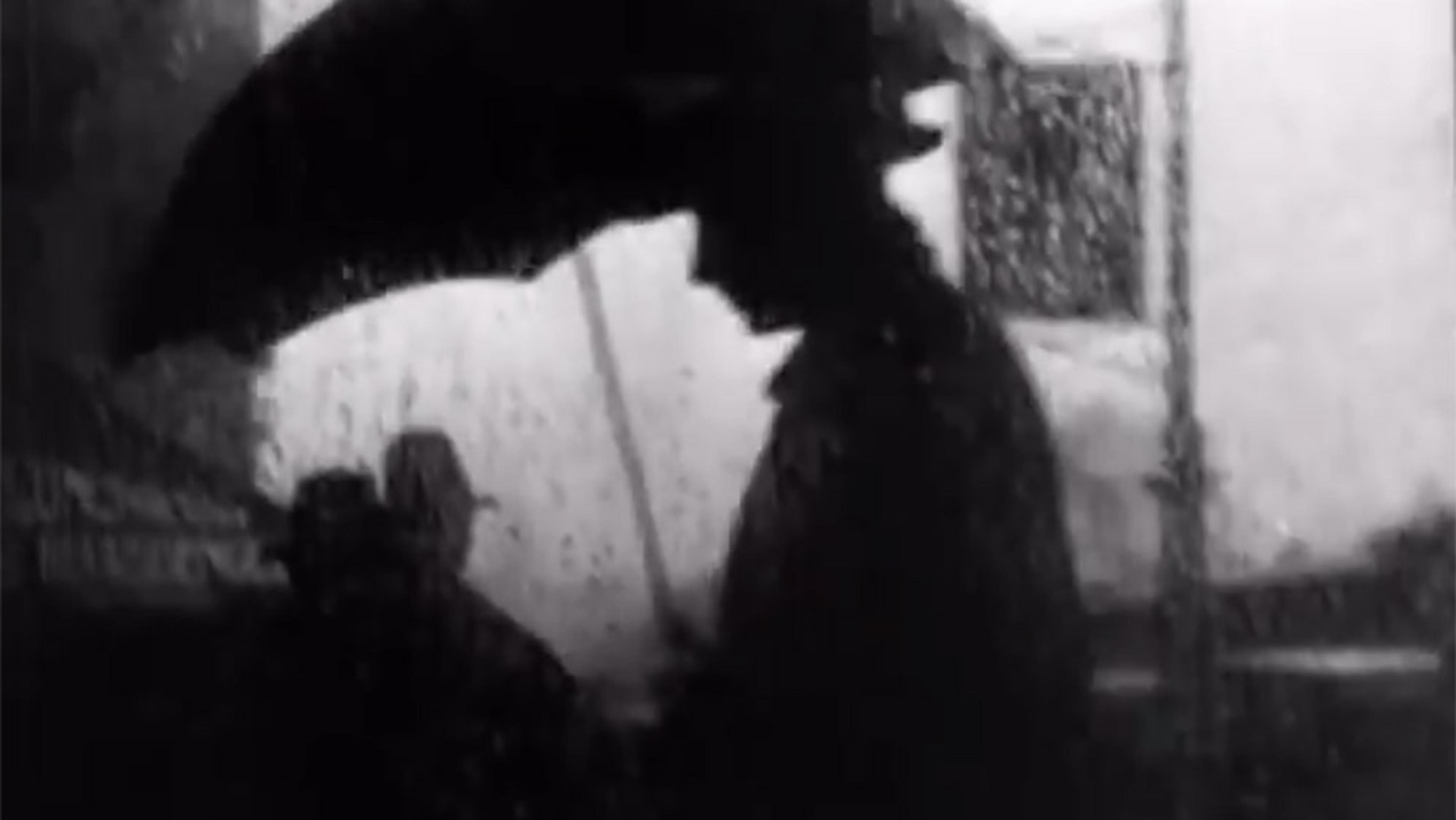 Black and white photo of a silhouetted person holding an umbrella in the rain, seen in profile against a blurry city background.