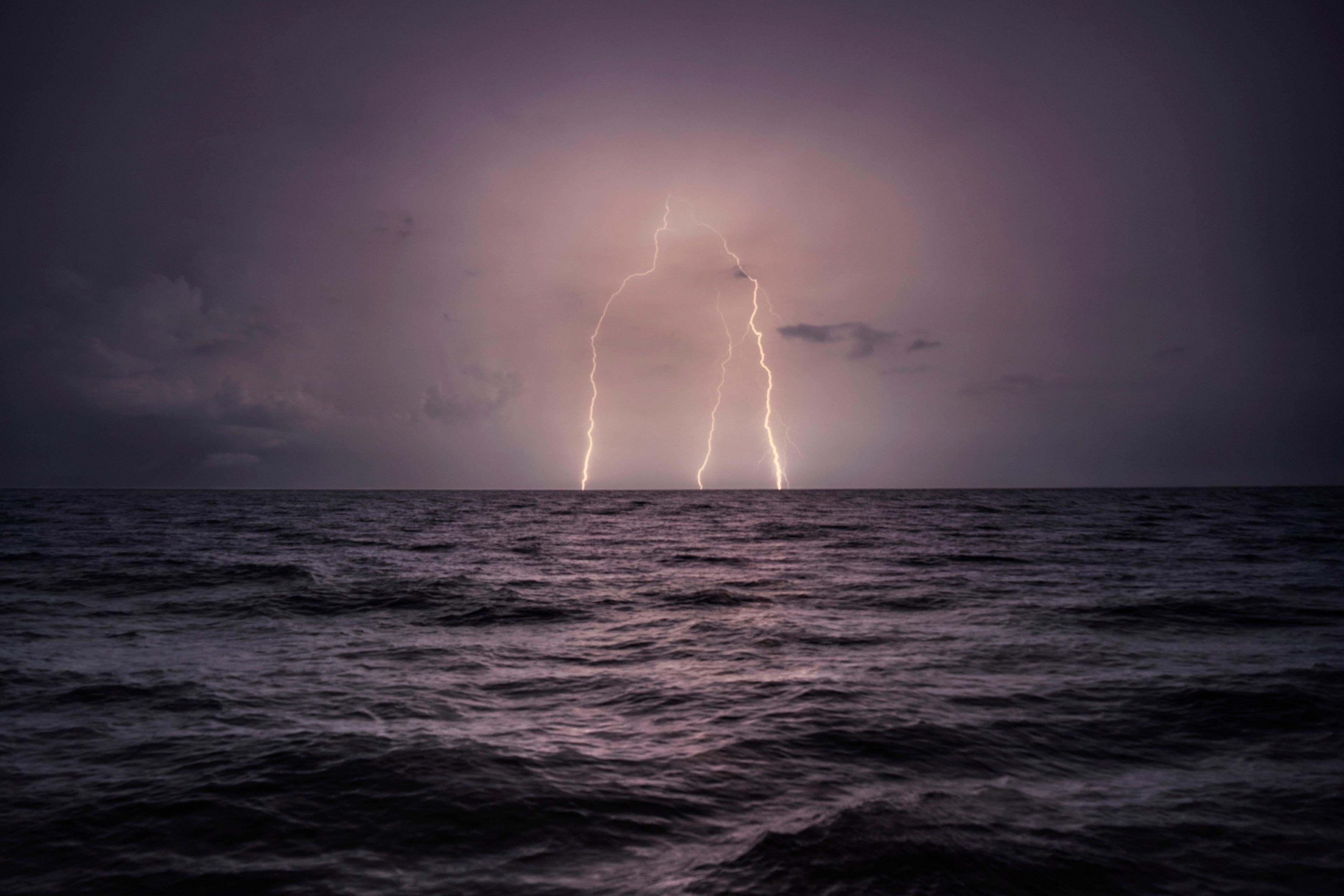 Photo of a dark ocean at night with two lightning strikes illuminating the cloudy sky.