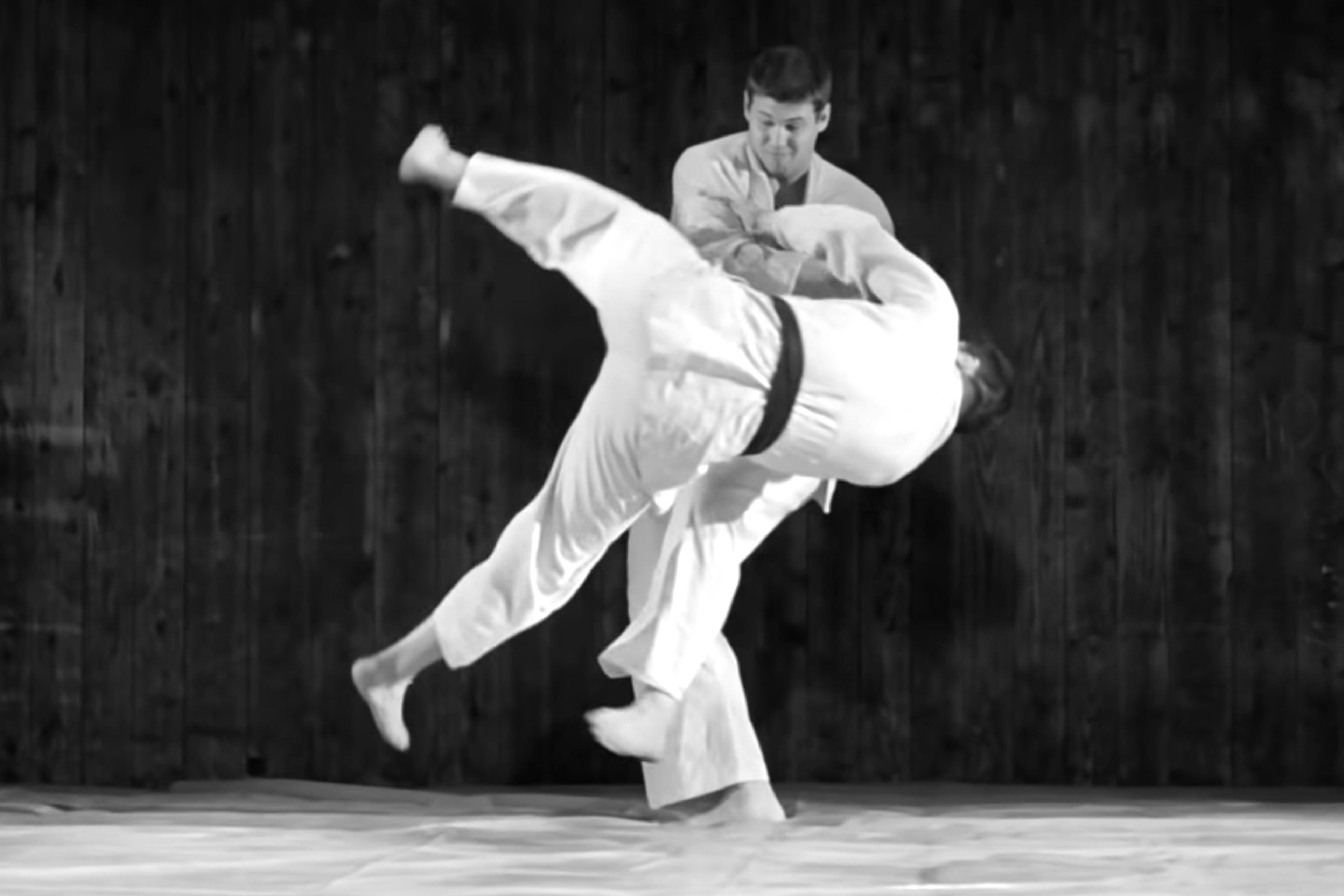 Black-and-white photo of two judokas; one is throwing the other who is airborne with legs up, set against a wooden background.