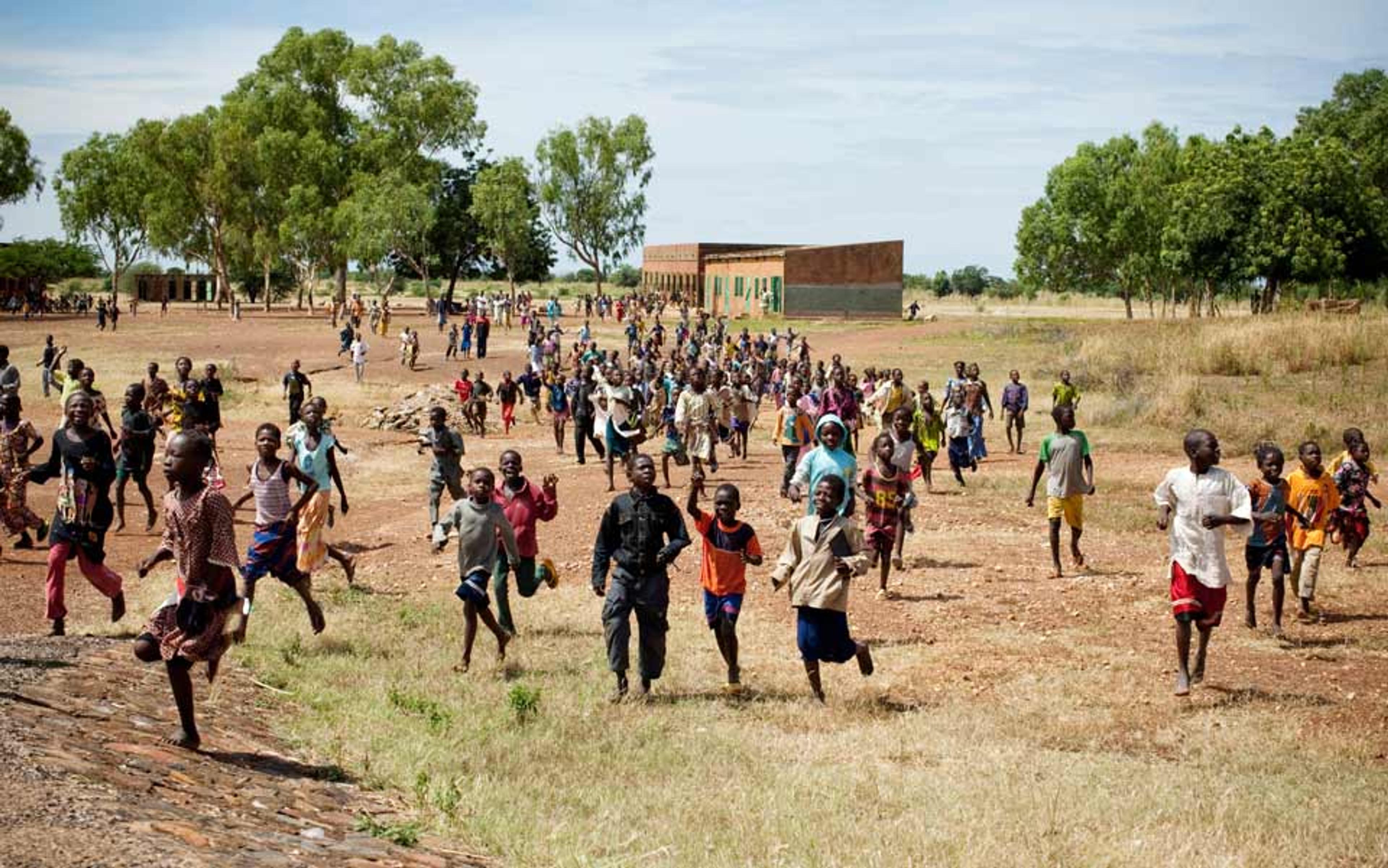 A large group of children running across an open field towards the camera with trees and a building in the background.